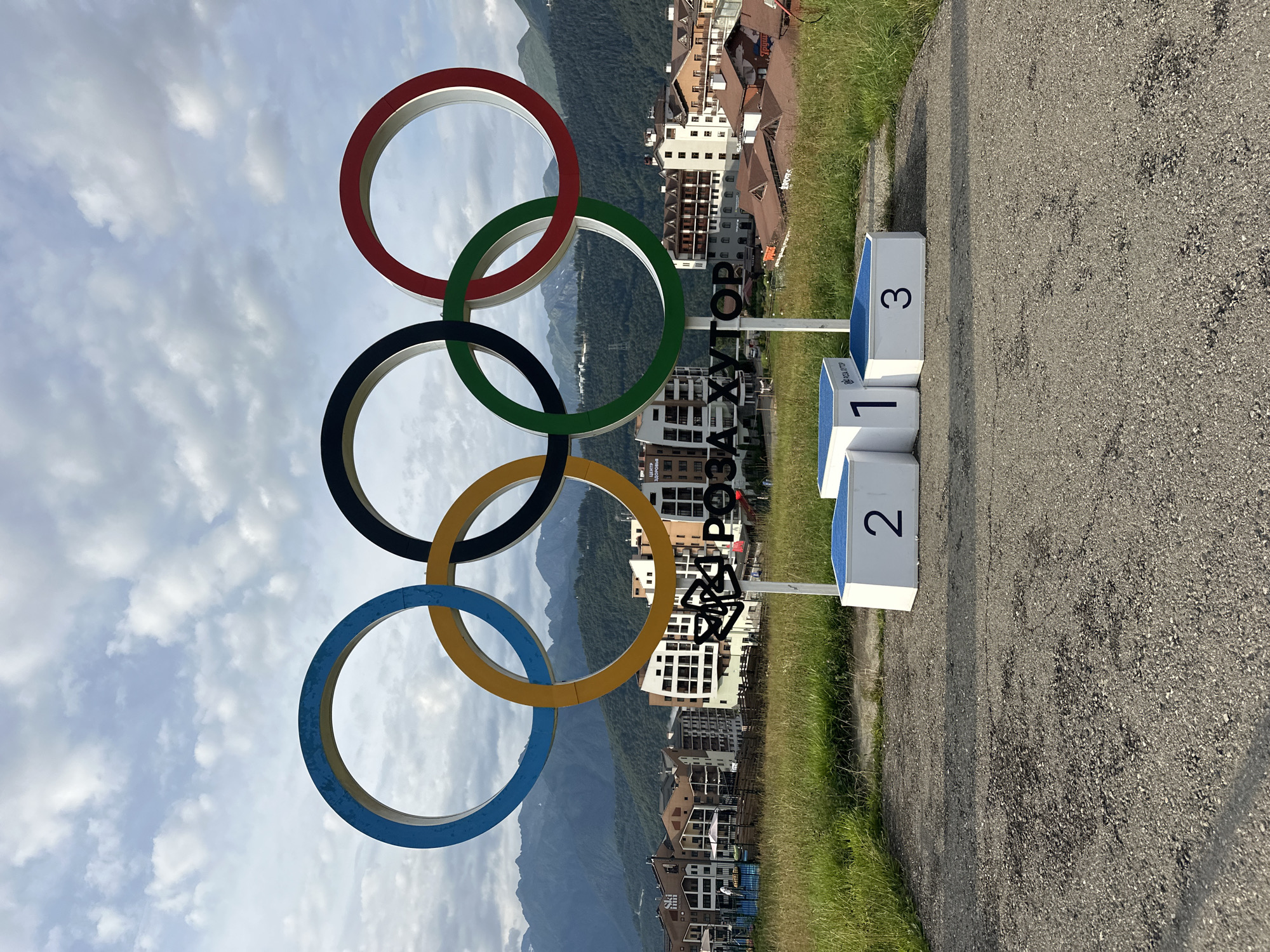 The image shows the Olympic rings installed in Estosadok, Russia. The rings are large, colorful, and mounted on a pole against a backdrop of mountains and buildings. In the foreground, there are three podiums numbered 1, 2, and 3. The podiums are white with blue tops. The Olympic rings are positioned behind the podiums. The Olympic rings are a prominent symbol of the Olympic Games and are often used in sports events. The scene appears to be a memorial or a tourist attraction, commemorating the Olympic Games held in Sochi in 2014. The presence of mountains and the architecture of the buildings suggest a ski resort or a similar mountainous region. The sky is cloudy, with patches of blue visible. The overall impression is one of a scenic and commemorative location.