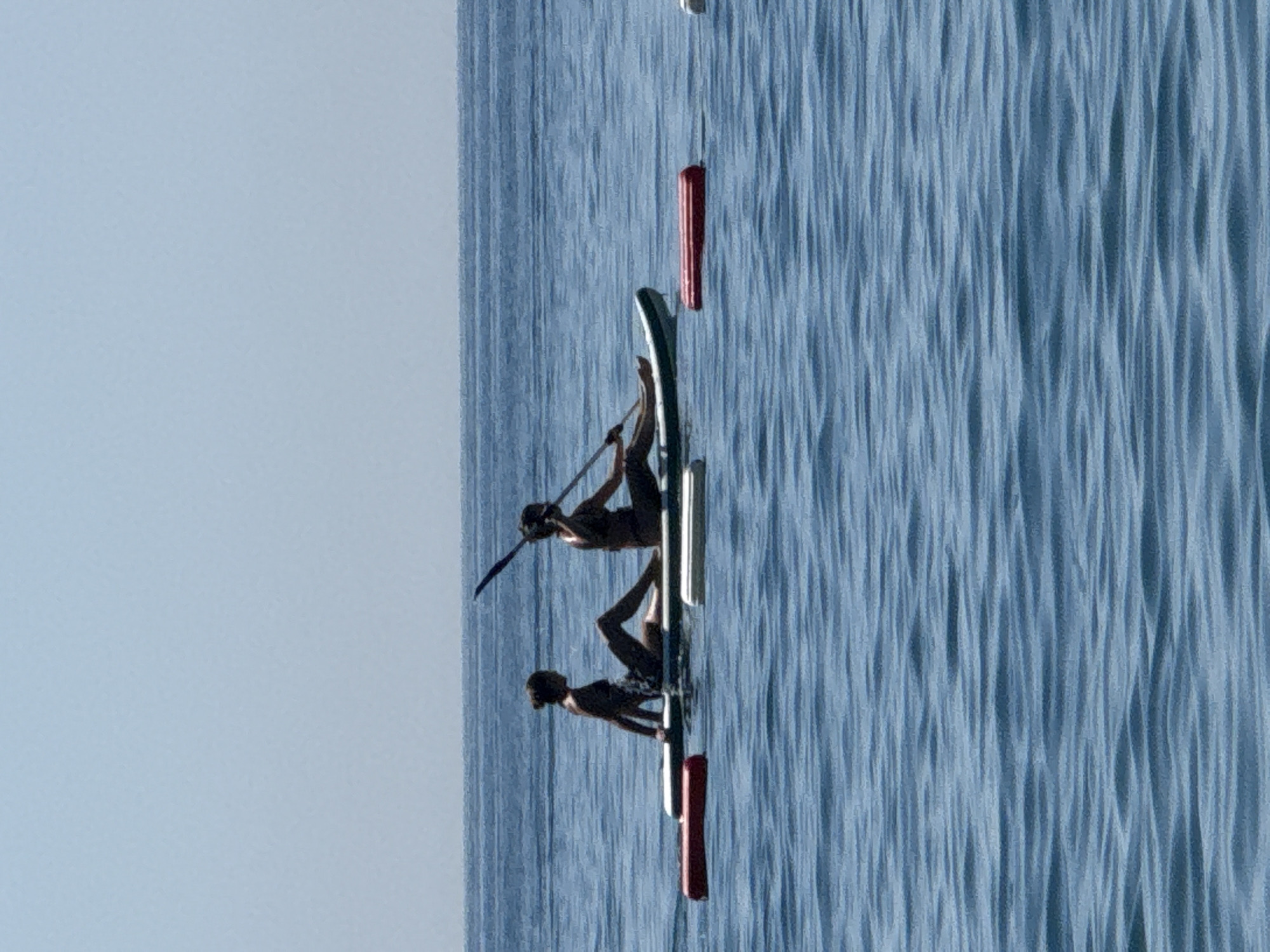 The image depicts two individuals engaged in stand-up paddleboarding on the tranquil, blue waters of Boreti, Montenegro. The two people are positioned side-by-side on separate paddleboards, with one individual holding a paddle. Both are seated on their respective boards. In the foreground, two red inflatable buoys are visible, floating on the water. The sky above is a clear, light blue, with the horizon line clearly separating the sea from the sky. The water is calm, with subtle ripples indicating a gentle movement on the surface.