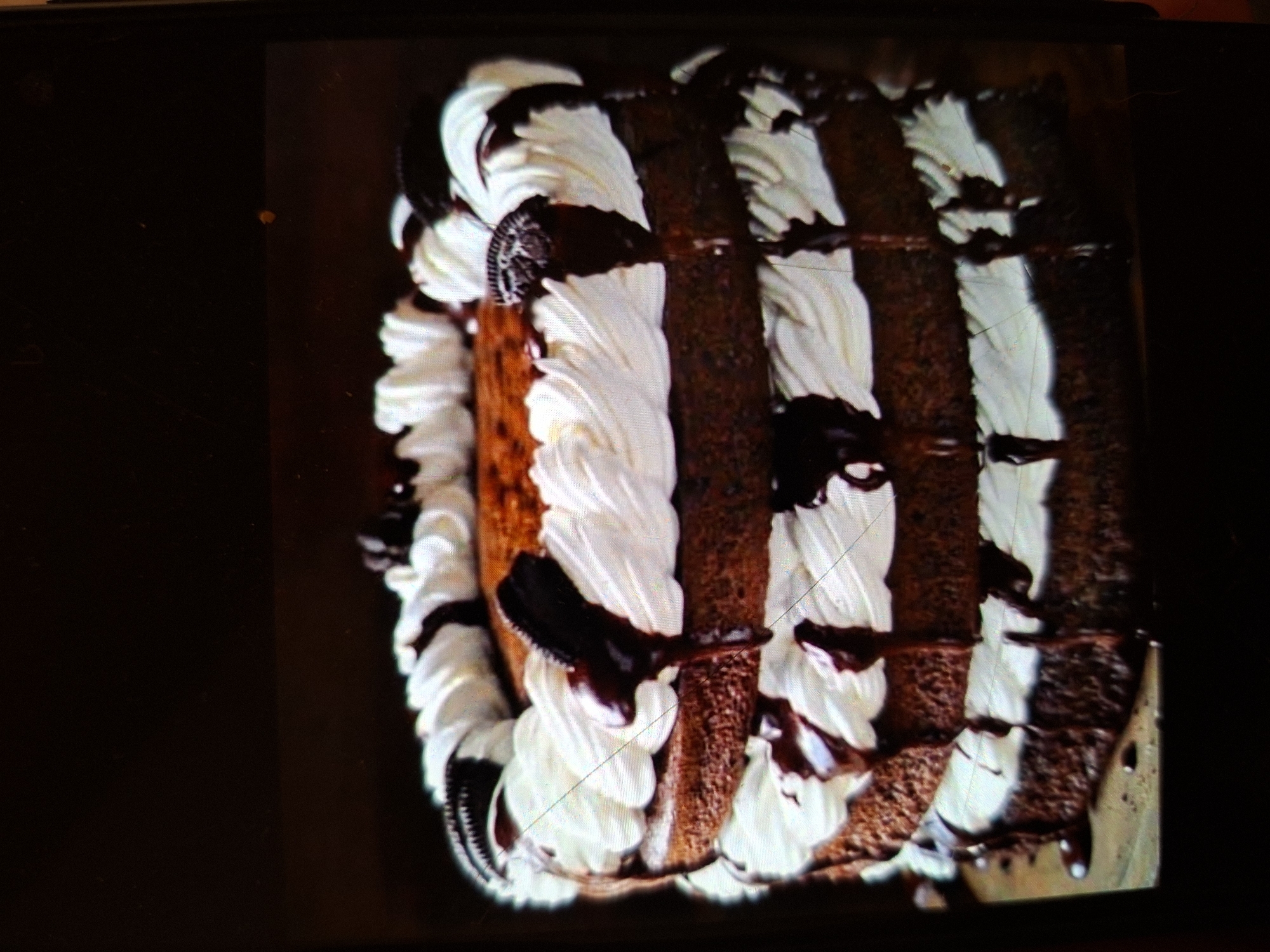 This is a close-up, low-angle, indoor shot of a three-layer chocolate cake adorned with white frosting and drizzles of chocolate sauce. Several Oreo cookies are placed on top and between the layers. The cake is positioned on a wooden surface against a dark background. The image is slightly blurry and has some glare. There is no indication of the city of Medvezhyi Ozyora, Russia, or any people, actions, or interactions.