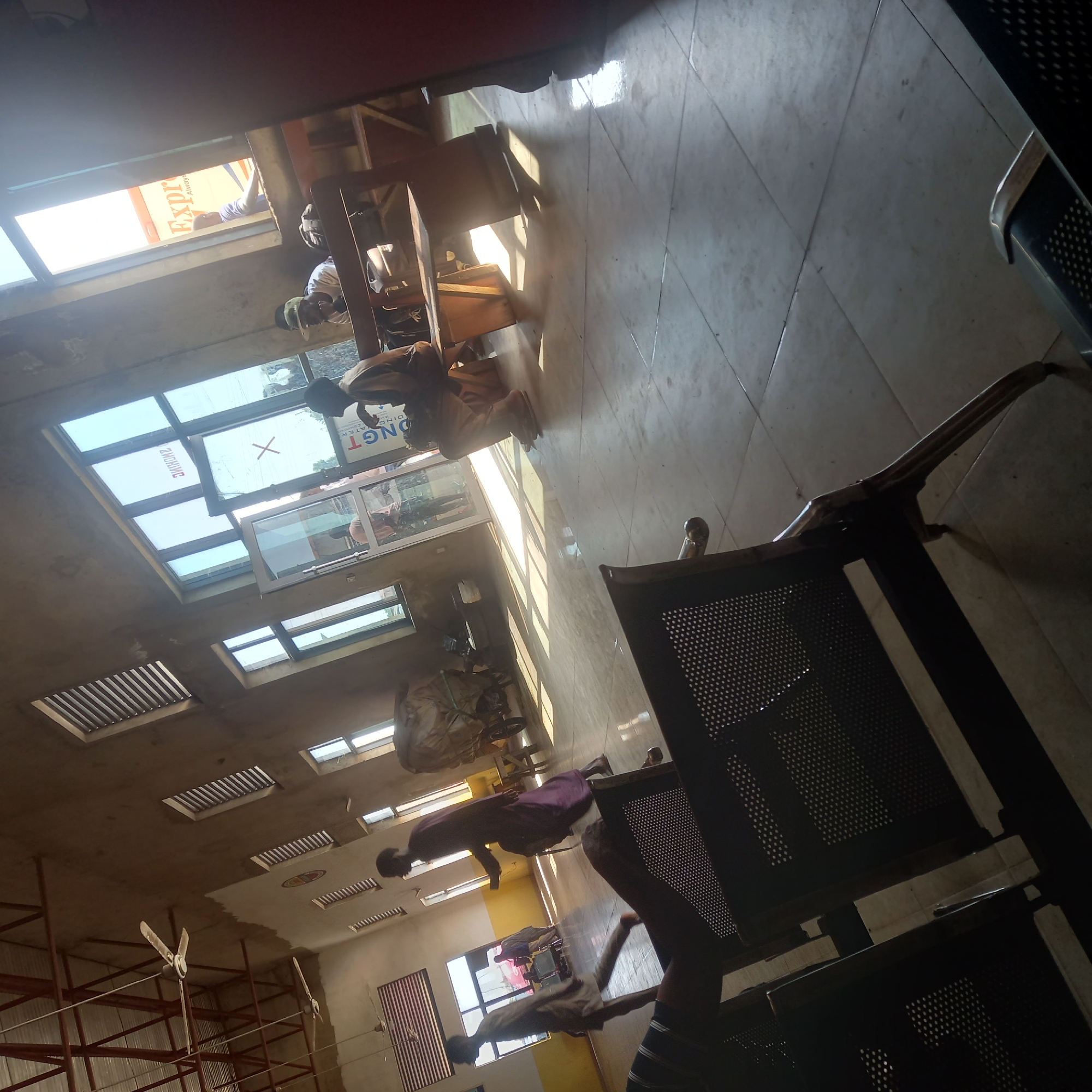 The image shows the interior of a waiting area, likely a bus station or similar transport hub, in Hayin Banki, Nigeria. The space has a tiled floor and concrete walls, with large windows providing natural light. Several ceiling fans are visible, indicating a warm climate. In the foreground, there are dark metal benches with perforated seats.
Several individuals are present in the scene. In the mid-ground, a person in a purple garment walks towards the left side of the frame. To their right, a person wearing a hat and white shirt sits on a bench, with a bag next to them. Further in the background, a person in a brown garment sits on a bench near a window. Through the windows, a truck or vehicle with the word "Express" visible is partially seen.
Other objects in the scene include a cart with a large bundle covered in a green tarp, and various bags and luggage. The overall impression is of a functional, somewhat rustic waiting area. The lighting suggests it is daytime, with strong shadows cast by the windows. The architectural style and the presence of the truck with "Express" branding suggest this is a commercial transport hub.