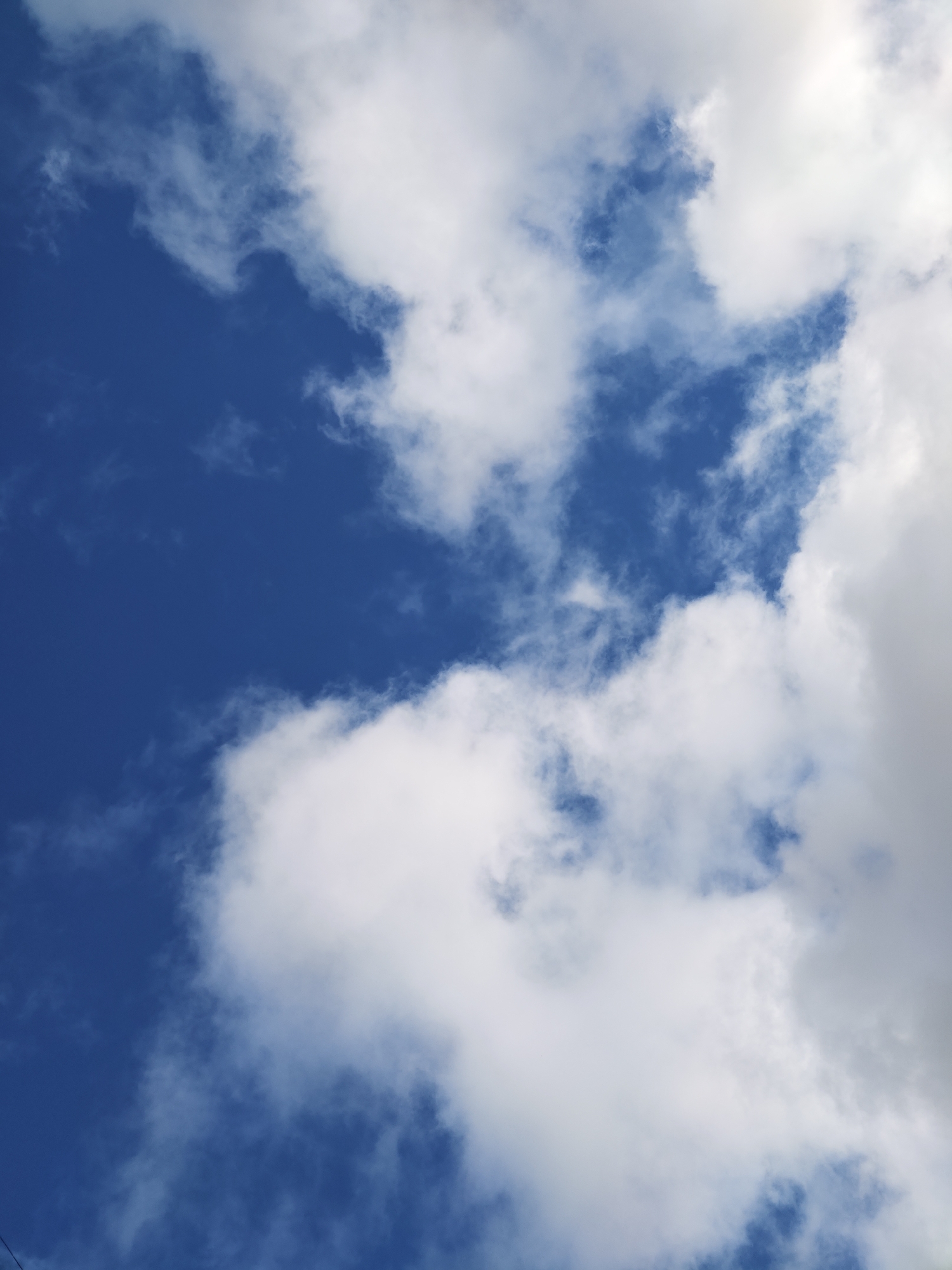 The image displays a vibrant blue sky partially obscured by white and greyish-white clouds. The scene is dominated by the dynamic interplay between patches of clear, deep blue sky and various cloud formations. On the right side, a substantial, bright white cloud mass stretches vertically, exhibiting depth and internal shadows that suggest its volume and thickness. Smaller, more fragmented, and wispy clouds are scattered across the upper left and central areas, appearing delicate against the vivid blue background. These clouds show soft edges and varying translucency, indicating different densities.
The lighting strongly suggests it is daytime, likely mid-morning or afternoon, as the clouds are brightly illuminated from above, enhancing their texture and form. The weather appears fair, characterized by scattered to broken cloud cover rather than a uniform overcast sky. No specific human activity or man-made structures are discernible within the frame, nor are there any unique natural landmarks that would visually confirm a particular geographical location. A very faint, thin, dark line is barely visible at the bottom left corner, but its identity is unclear.
The provided context specifies the location as Chaikovsky, Russia, though this cannot be independently verified from the visual information alone, as the image presents a generic sky view. There is no visible text present in the image. The overall impression conveyed is one of natural beauty, expansiveness, and the serene quality of a bright, cloudy day.