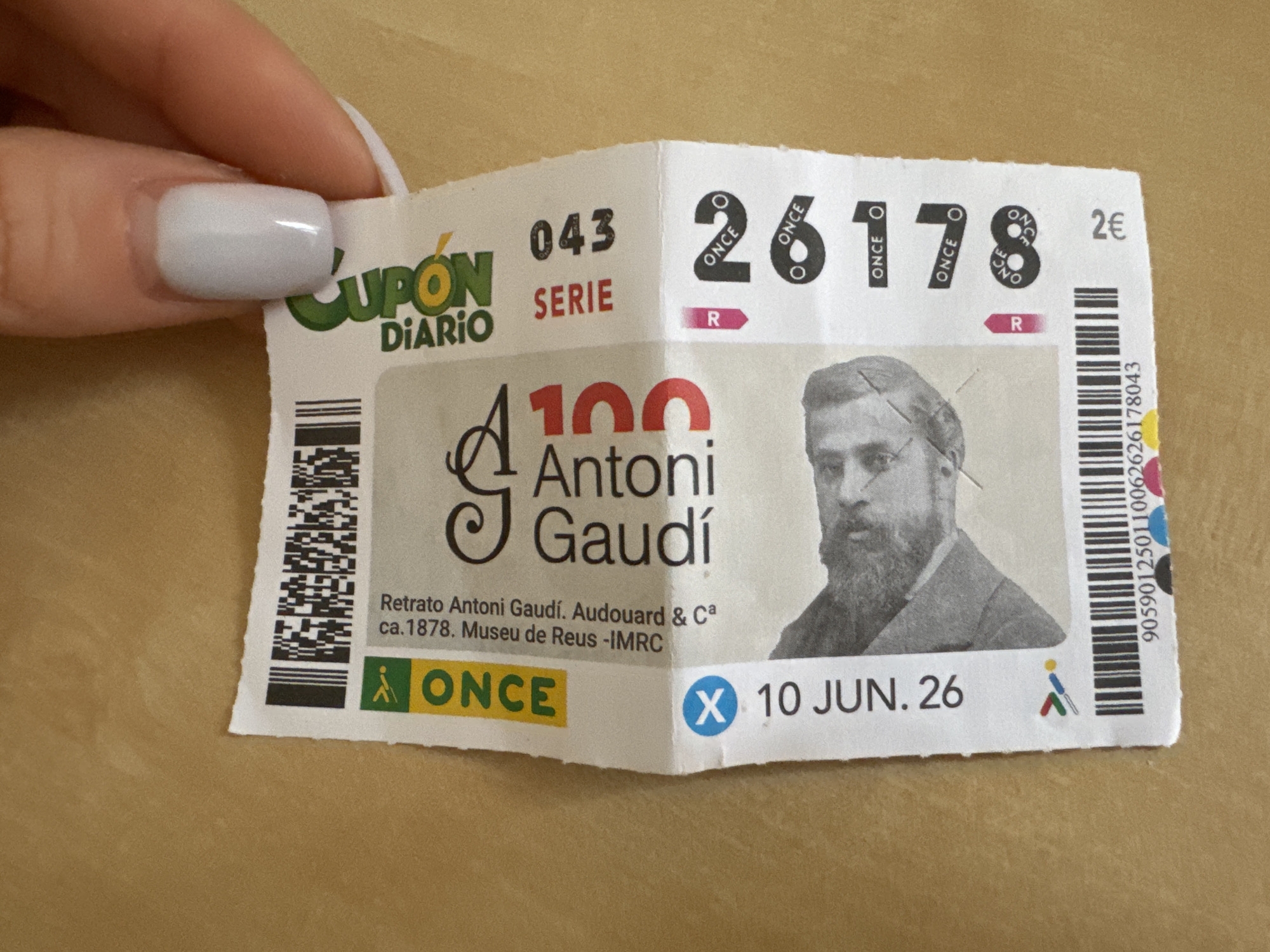 The image shows a hand holding a lottery ticket. The ticket is white with text and graphics printed on it. The text includes "ONCE", "CUPÓN DIARIO", "SERIE 043", "10 JUN. 26", and numbers "100", "26178". There is also a portrait of a man on the ticket. The background is a textured surface, possibly a table. The hand is holding the ticket from the top right corner, with fingernails painted a light blue color. The location in the city of Murcia, Spain is not ascertainable from the image itself.