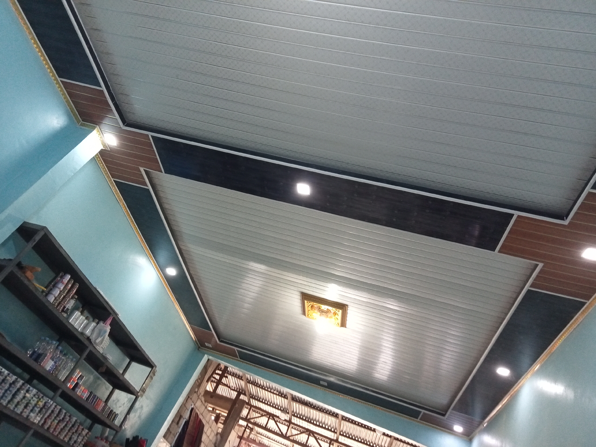 This is a low-angle interior shot of a room with a decorative, paneled ceiling. The ceiling is made of white, horizontal planks and is bordered by dark gray and wood-patterned panels. Several recessed lights are visible on the ceiling. A single ornate light fixture with a yellowish glow is centrally located in one section. To the left, a turquoise wall meets the ceiling, and a black, multi-tiered shelf unit is mounted on the wall, filled with various bottles and containers. The room appears to be a residential or small commercial space in Melayu Besar Kota, Indonesia. The overall impression is one of a renovated or newly decorated interior.