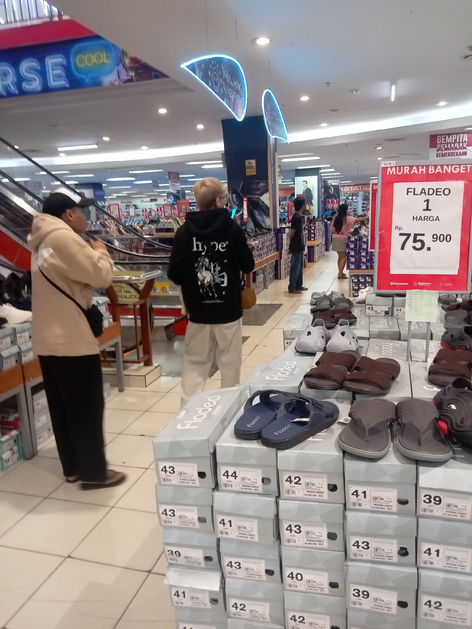 The image shows a retail store interior, likely a shoe store, with stacks of shoe boxes and footwear displayed. In the foreground, there are several pairs of sandals and flip-flops, some displayed on top of boxes and others laid out on the floor. The shoe boxes are stacked neatly, with labels indicating sizes and possibly product codes and prices. A large red and white sign with "MURAH BANGET" (meaning "Very Cheap") is prominent, advertising a product called "FLADEO 1" for Rp. 75,900.
In the mid-ground, two individuals are visible. On the left, a person wearing a light brown hoodie and dark pants with a crossbody bag is looking to the right. On the right, a person in a black hoodie with white lettering and light-colored pants is facing away from the camera. In the background, more shelves and displays of shoes are visible, along with escalators and signage. One sign in the distance appears to read "GEMPITA SEMARAK KEMERDEKAAN" (meaning "Festive Glorious Independence").
The environment appears to be a well-lit, modern retail space with tiled flooring. The overall impression is that of a busy shopping area offering discounted footwear. Based on the style of the store, the signage in Indonesian, and common retail chains in the region, this scene is consistent with a department store or a large footwear retailer located in Batam, Indonesia, such as Robinson Department Store.