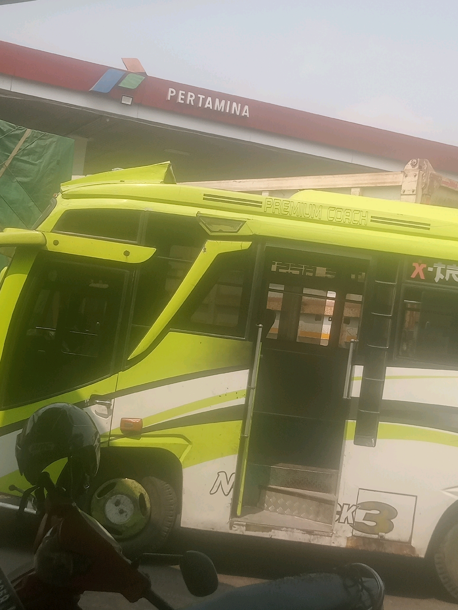In Pendawa, Indonesia, a lime green bus with "PREMIUM COACH" and "X-TR" written on its side is parked at a Pertamina gas station. The bus has its doors open, revealing stairs leading into the interior. A motorcycle with a dark helmet is parked in front of the bus. The sky is overcast.