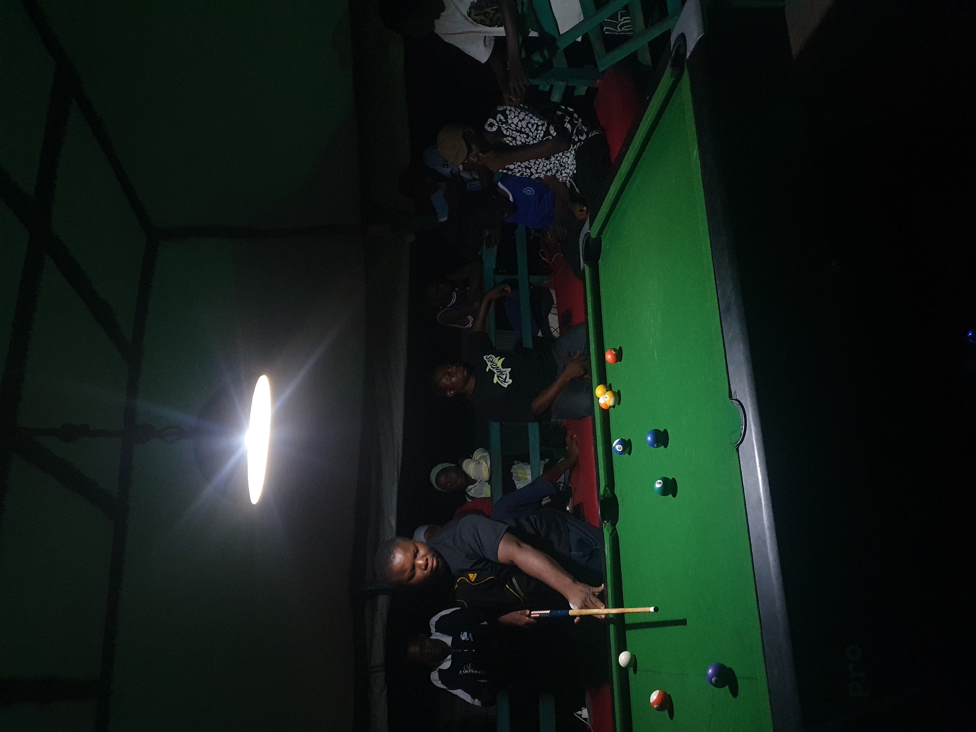 The image depicts a group of people gathered around a pool table in an indoor setting, possibly a bar or a recreational space, at night. The lighting is dim, with a bright spotlight illuminating a portion of the scene, casting long shadows and creating a dramatic effect.
Several individuals are visible, some standing and observing, while others are actively engaged with the pool table. One man in the foreground, wearing a dark shirt, is holding a pool cue and appears to be preparing to take a shot. Another person is seated at the table, watching the game. The pool table itself is green, with several colored balls scattered across its surface, including a red ball, blue balls, and a yellow-striped ball.
The background reveals a dimly lit room with what appear to be wooden structures and seating. The overall atmosphere suggests a social gathering or a casual game night. The image is oriented such that the pool table runs diagonally across the frame. The location is identified as Lokoja, Nigeria, and the time is clearly nighttime due to the artificial lighting and the darkness outside. No specific weather conditions are discernible. There is no visible text in the image.