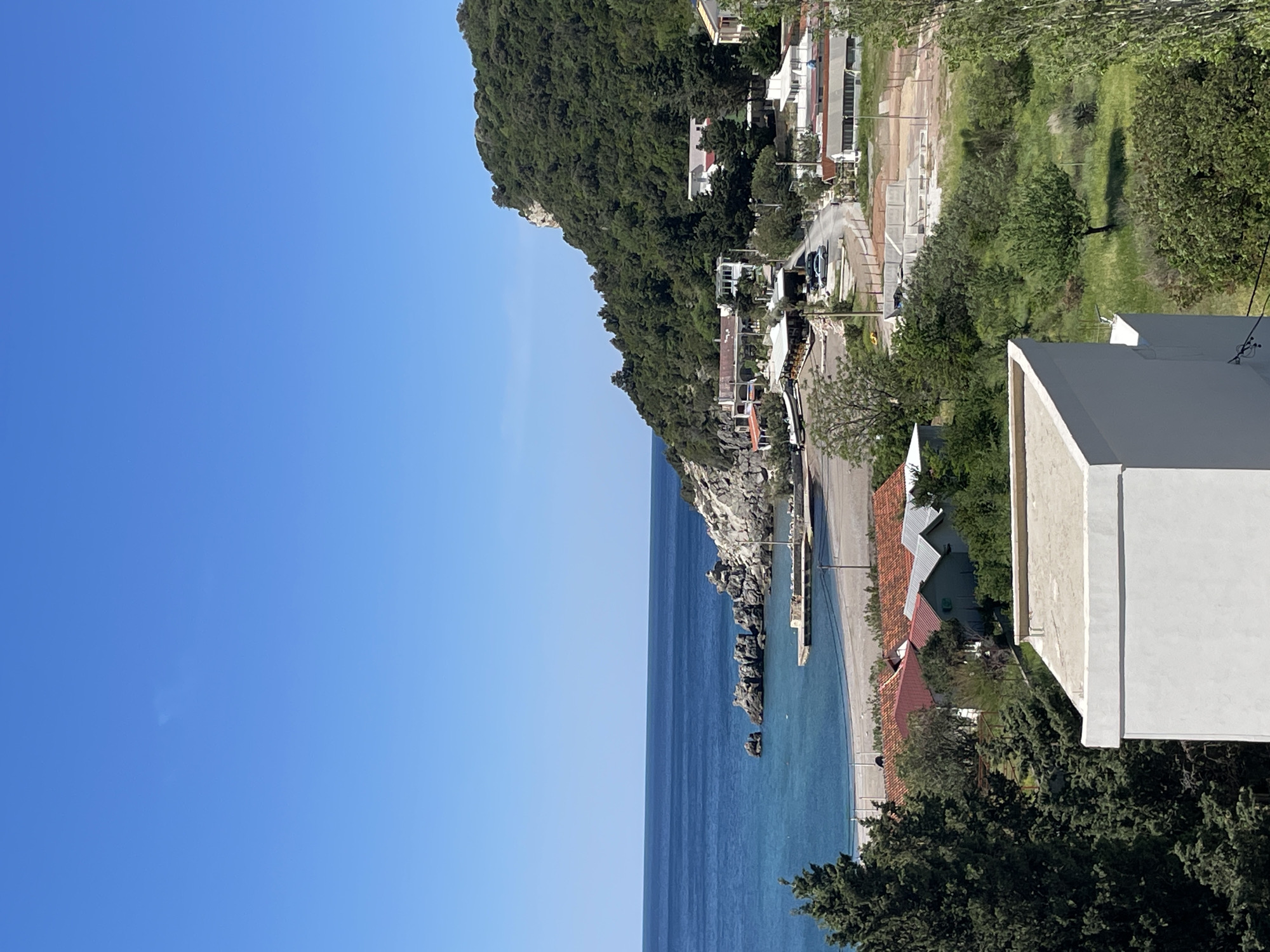 This image presents a vibrant coastal landscape in Čanj, Montenegro, viewed from an elevated perspective. The scene is dominated by a clear, azure sky and the tranquil blue waters of the Adriatic Sea, stretching horizontally across the middle and left portion of the frame. The sea meets a rocky coastline and a narrow, light-colored beach. A small pier or jetty extends into the water from the beach area.
To the right, a steep, heavily forested hillside rises, covered in lush green trees. Along the base of this hill, and extending further inland, is a settlement with various buildings. These structures include houses with red-tiled roofs, as well as some modern, multi-story buildings with flat roofs and white or light-colored facades. A road or path winds along the coastline, with a few parked cars visible. Further back, beyond the immediate developed area, patches of grassy terrain and more trees are seen.
The time of day appears to be sunny daylight, likely mid-morning or afternoon, indicated by the bright illumination and distinct shadows cast by trees and buildings. The weather is clear and calm. No specific activities or people are discernible within the frame, emphasizing the natural beauty and architectural elements of the coastal setting. There is no visible text in the image.