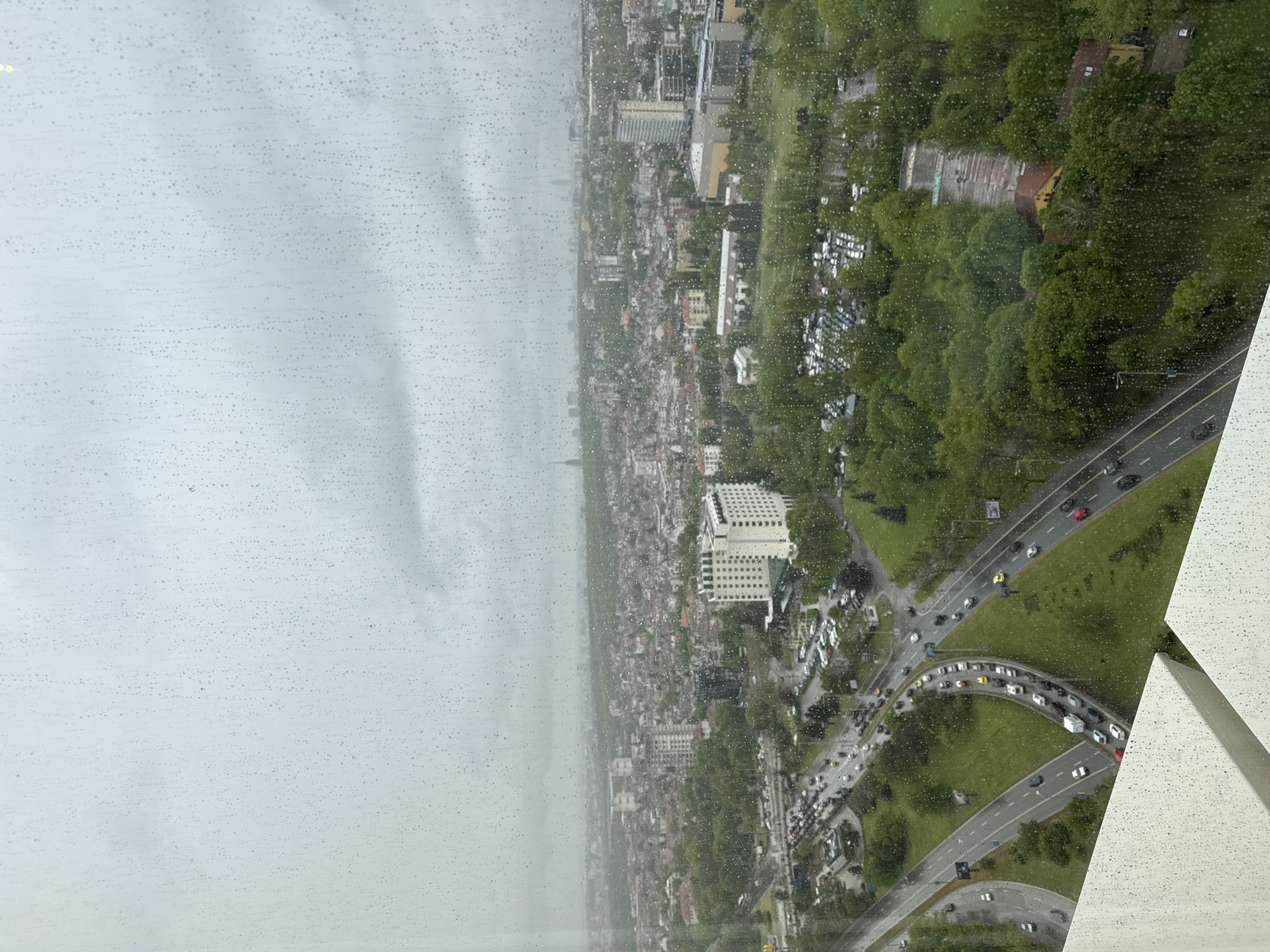 This image presents a high-angle, panoramic view of a sprawling urban landscape, likely Sofia, Bulgaria, as seen through a window covered in numerous rain or water droplets. The overall impression is that of a cloudy, overcast, and rainy day.
The left side of the frame is dominated by a light grey, hazy sky, heavily obscured by water droplets on the glass, which extend across most of the view. Towards the horizon, the city gradually fades into the atmospheric haze.
The right side of the image offers a more detailed perspective of the cityscape. A dense mix of buildings, ranging from multi-story structures to smaller, more traditional architecture, populates the area. A prominent, large, light-colored building with a grid-like pattern of windows stands out in the midground, possibly a commercial or hotel building. Interspersed throughout the urban fabric are substantial green areas, including parks and tree-lined expanses, providing a natural contrast to the built environment.
A network of roads with active traffic is visible in the foreground and midground. Cars, some appearing to be in queues, navigate the wet-looking asphalt, particularly at a complex intersection towards the bottom right. Several parking lots filled with vehicles are also visible. In the bottom right corner, a distinctive white, angular architectural element of the building from which the photograph was taken juts into the frame, also adorned with water droplets.
The time of day is clearly daytime, though the heavy cloud cover casts a soft, diffuse light over the entire scene. There are no clearly discernible people or specific events taking place, portraying a typical, bustling city environment amidst rainy weather. No visible text is present within the frame.