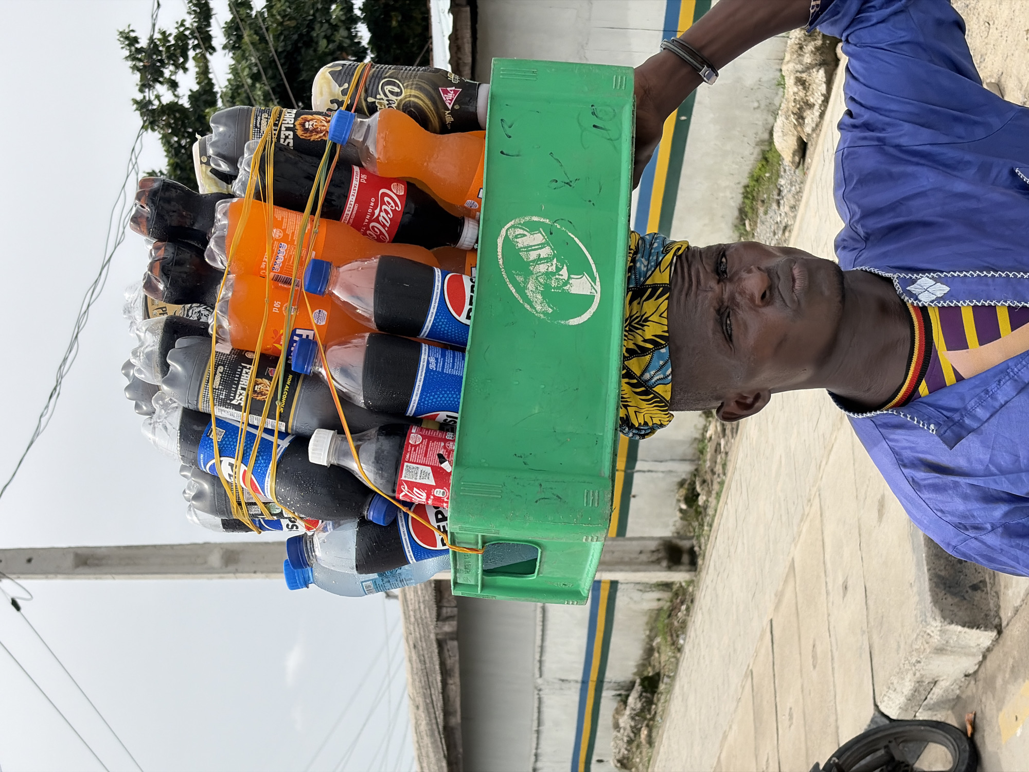 A man in Igboefon, Nigeria, carries a large stack of bottled beverages on his head. The bottles, including Coca-Cola, Pepsi, and Charles beverages, are secured with rubber bands and contained within a green plastic crate. He wears a colorful headwrap and a blue shirt. He walks on a paved surface. Power lines are visible in the background.