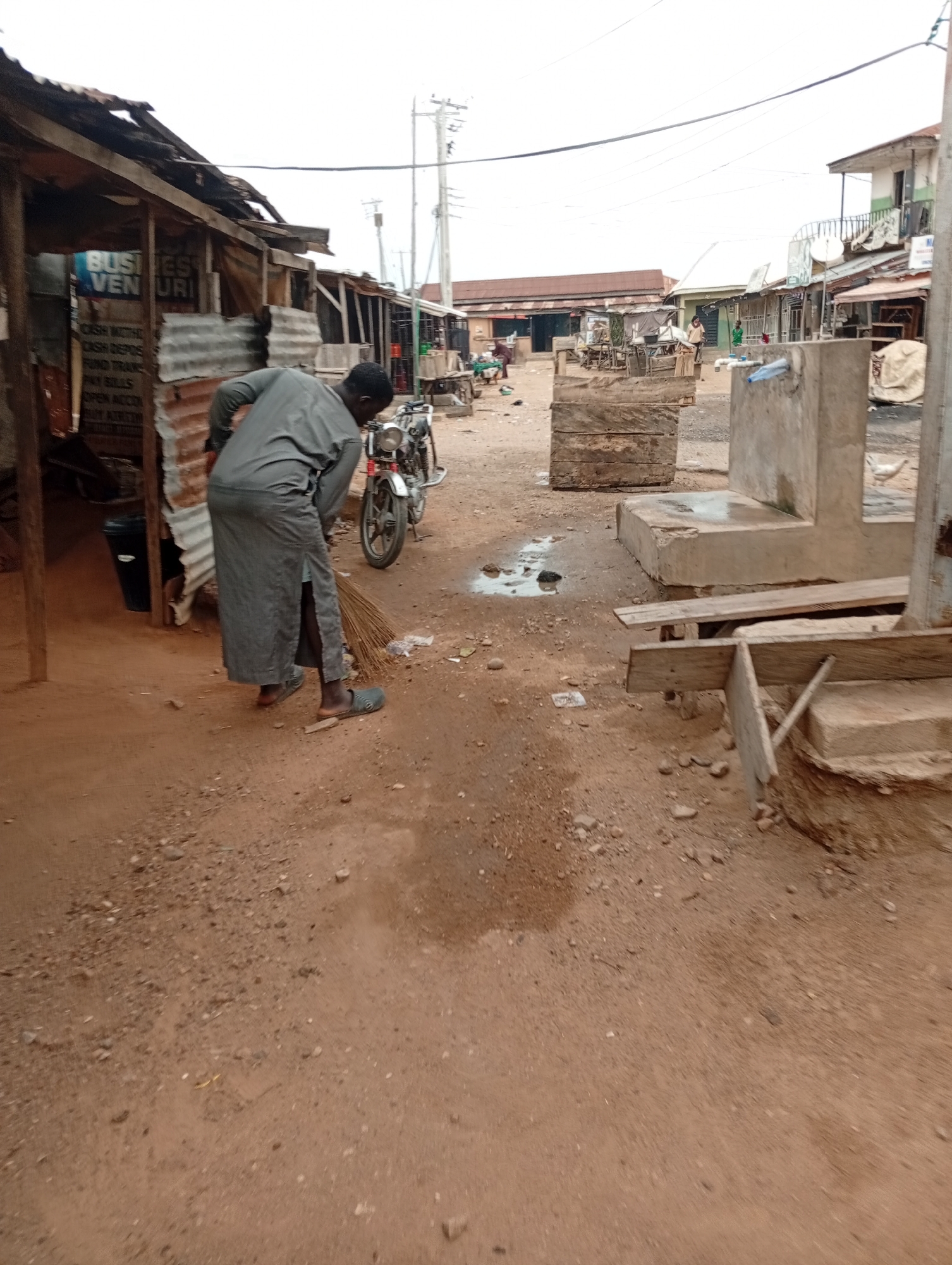 A man in a long gray tunic sweeps a dirt road in Zango-ndaji, Nigeria. The road is unpaved and cluttered with debris. He wears sandals. Behind him stands a motorcycle. To the left is a dilapidated structure with the words "BUSINESS VENURI" visible on a sign. Across the road are various makeshift stalls and buildings. The sky is overcast. In the distance, people are visible walking around. Concrete structures sit on the right side of the frame.