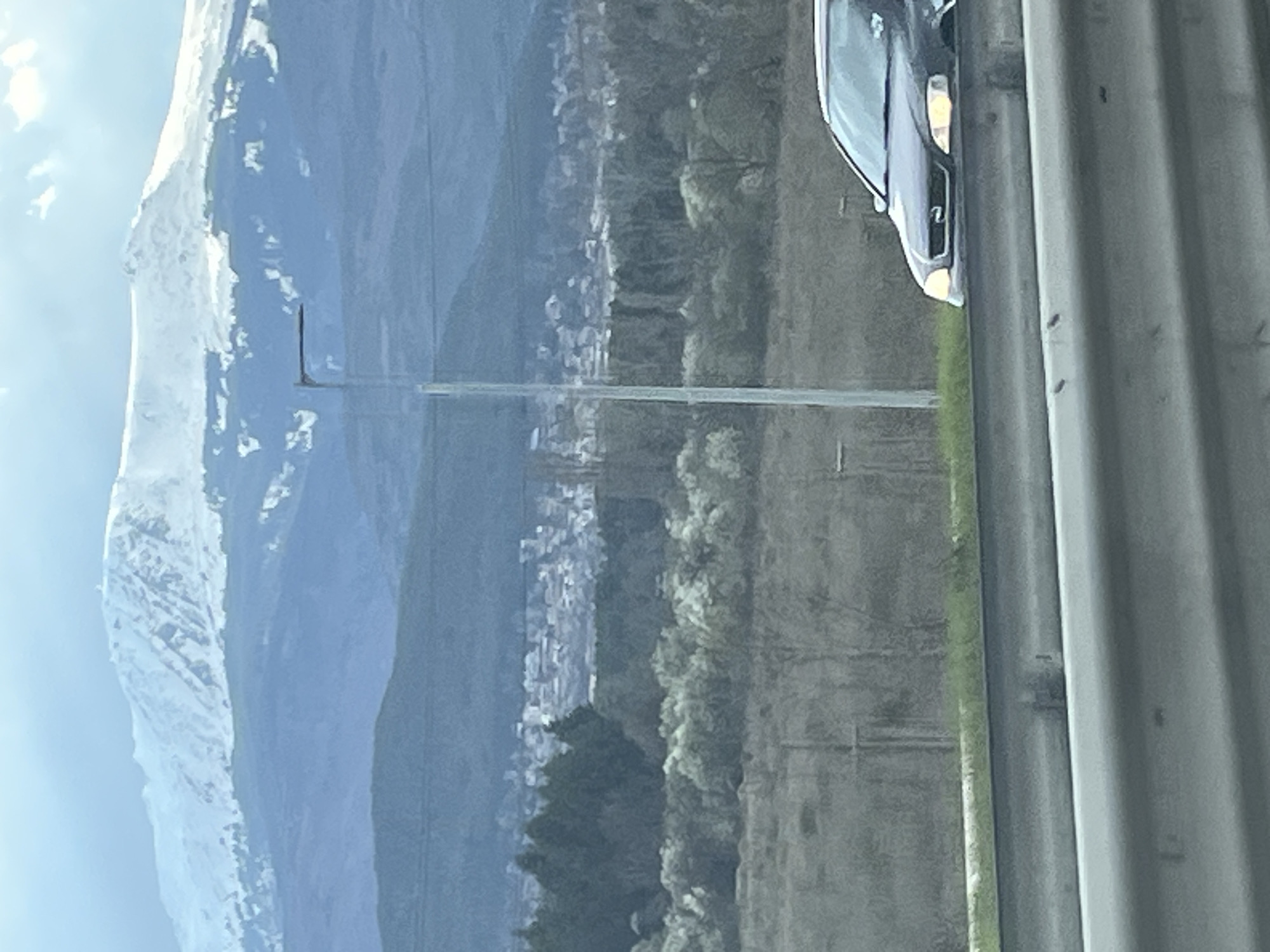 The image shows a view from a vehicle, likely a car. On the right side, there is a portion of a vehicle (headlights visible) and a highway divider. In the background, there is a landscape with mountains partially covered in snow, a town or city, trees, and utility poles. The location appears to be near a city with mountainous terrain.