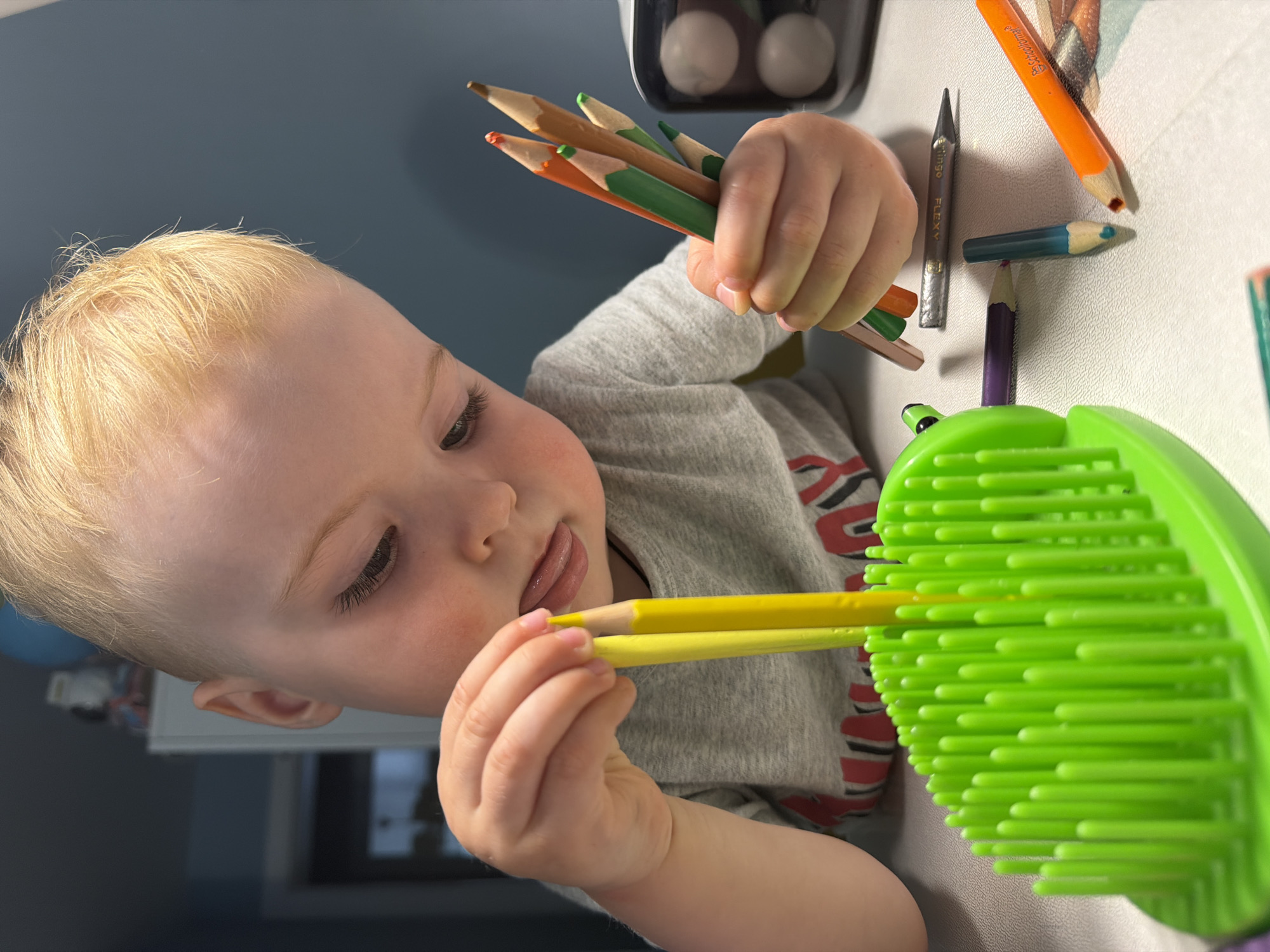 A close-up, slightly rotated, interior shot shows a fair-skinned toddler with blond hair holding and manipulating colored pencils over a green plastic object. The child is wearing a gray t-shirt with red and black markings. The toddler holds a cluster of pencils in one hand and inserts a yellow pencil into the green object with the other. Other colored pencils and a pencil sharpener are scattered on a white surface. A white object is visible in the background. There is no information available to identify the location as Votkinsk, Russia.