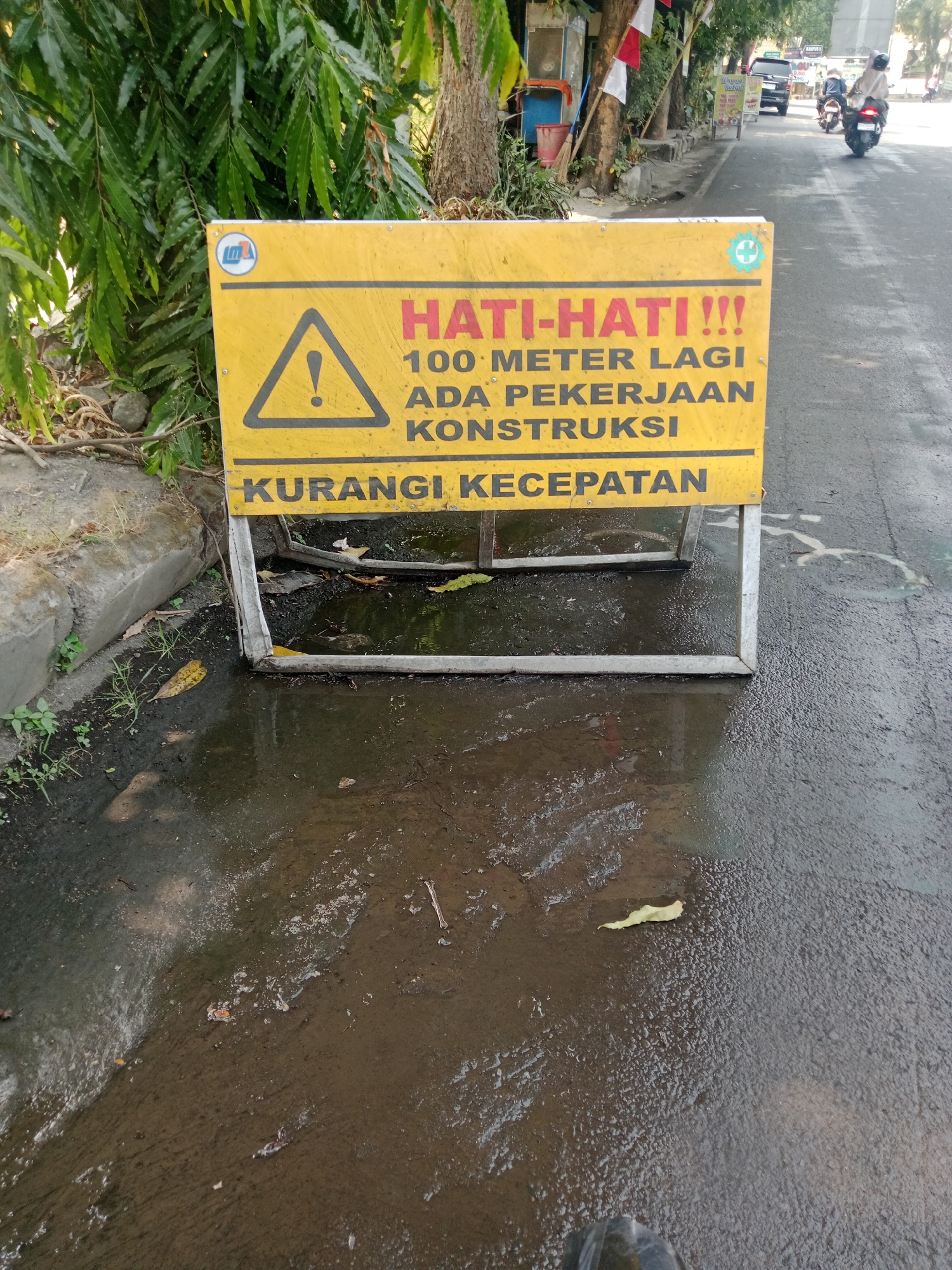 A yellow warning sign with black lettering is propped up on a metal stand on the side of a wet asphalt road. The sign reads "HATI-HATI!!!" (Be Careful!!!) in large red capital letters, followed by "100 METER LAGI ADA PEKERJAAN KONSTRUKSI" (100 meters ahead there is construction work) and "KURANGI KECEPATAN" (Reduce speed). A black triangle with a white exclamation mark is present on the left side of the sign. To the left of the sign, a concrete curb is visible, with some vegetation and leaves on the ground. The road is wet, with puddles of water and debris on the surface. In the background, on the right side of the image, a motorcyclist wearing a helmet and light-colored clothing is riding away from the camera. Further in the background, there are some buildings, trees, and other vehicles. The overall scene suggests a road in an urban or semi-urban area in Kediri City, Indonesia, where construction is taking place.