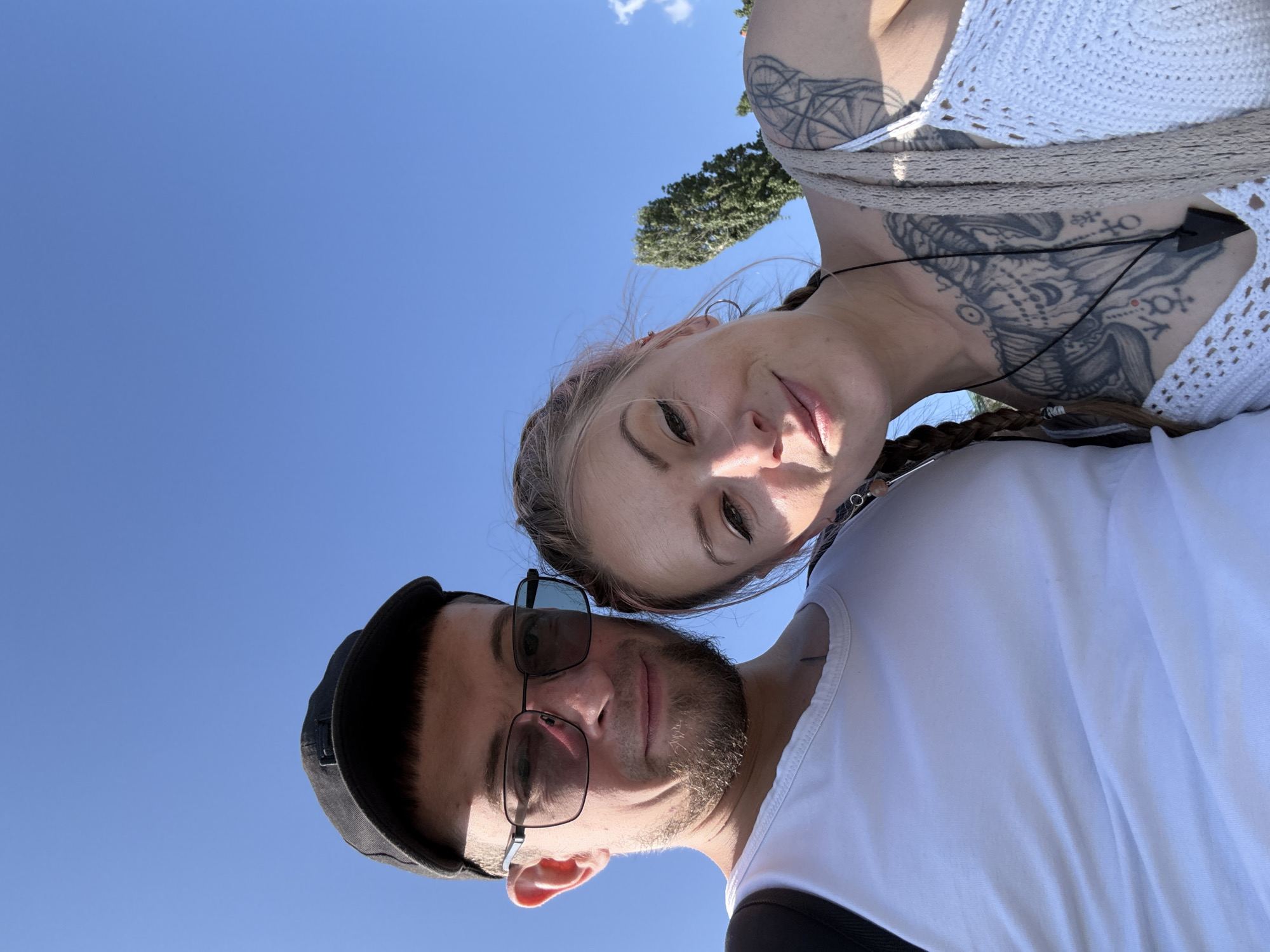 This is a selfie taken outdoors with a clear blue sky in the background. A man wearing a black baseball cap, sunglasses, and a white tank top is on the left. He has a beard and is looking towards the camera. Next to him, on the right, is a woman with blonde hair, some of which is pink. She is also looking at the camera, smiling. She has a braid and visible tattoos on her chest and shoulder, as well as a necklace with a pendant. A tall, green tree is visible in the background behind the woman. The overall impression is that of a couple enjoying a sunny day. The location is identified as Saratov, Russia.