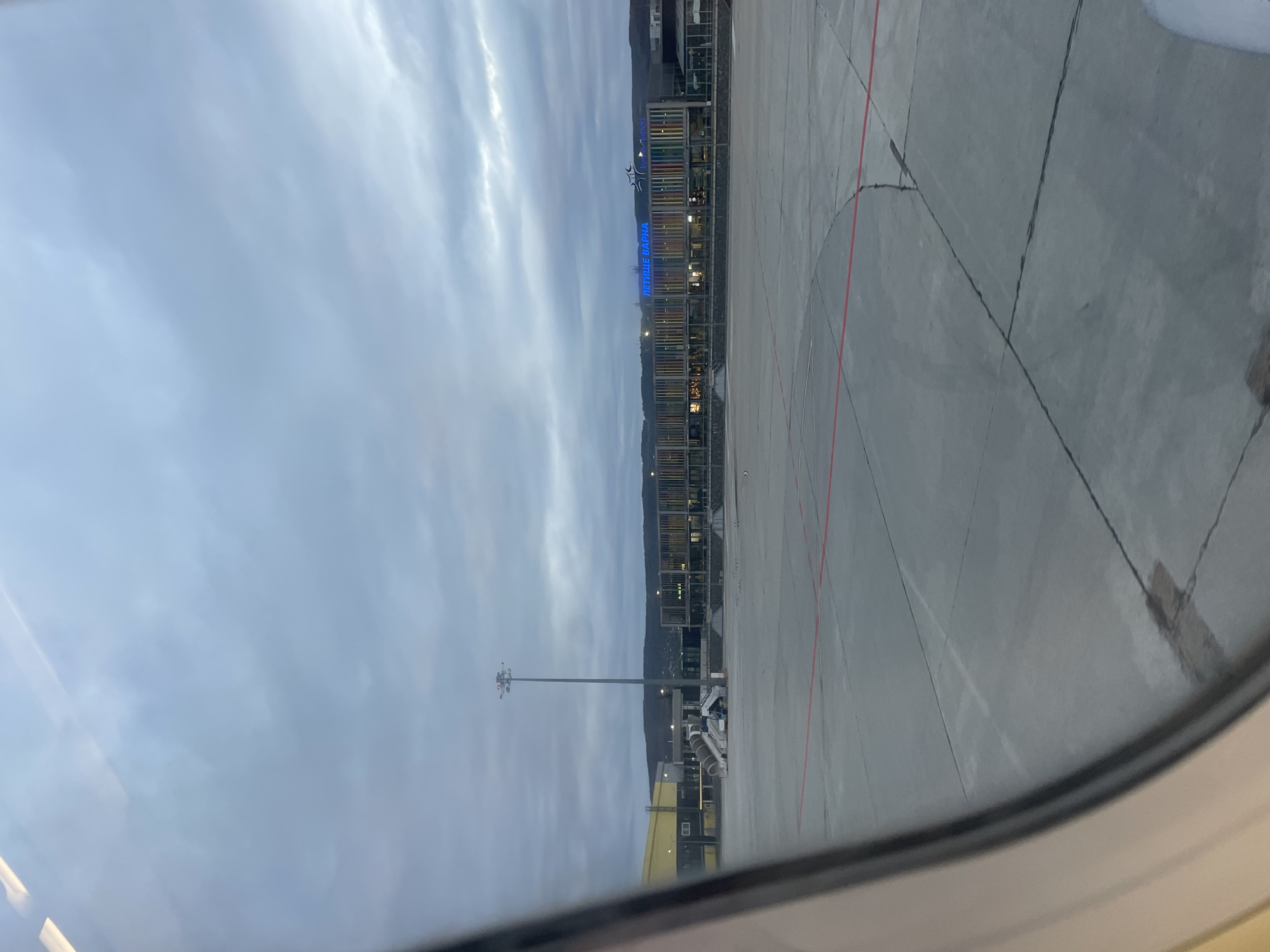The image shows a view from an airplane window looking out onto Varna Airport (VAR) in Aksakovo, Bulgaria. The airport terminal building is visible with its architectural facade and signage in Bulgarian (ЛЕТИЩЕ ВАРНА) spelled out in blue letters. Airport markings are visible on the concrete apron. A utility pole, possibly a lighting tower, is standing near some parked ground support equipment, perhaps a jet bridge or boarding stairs. The sky overhead is overcast.
