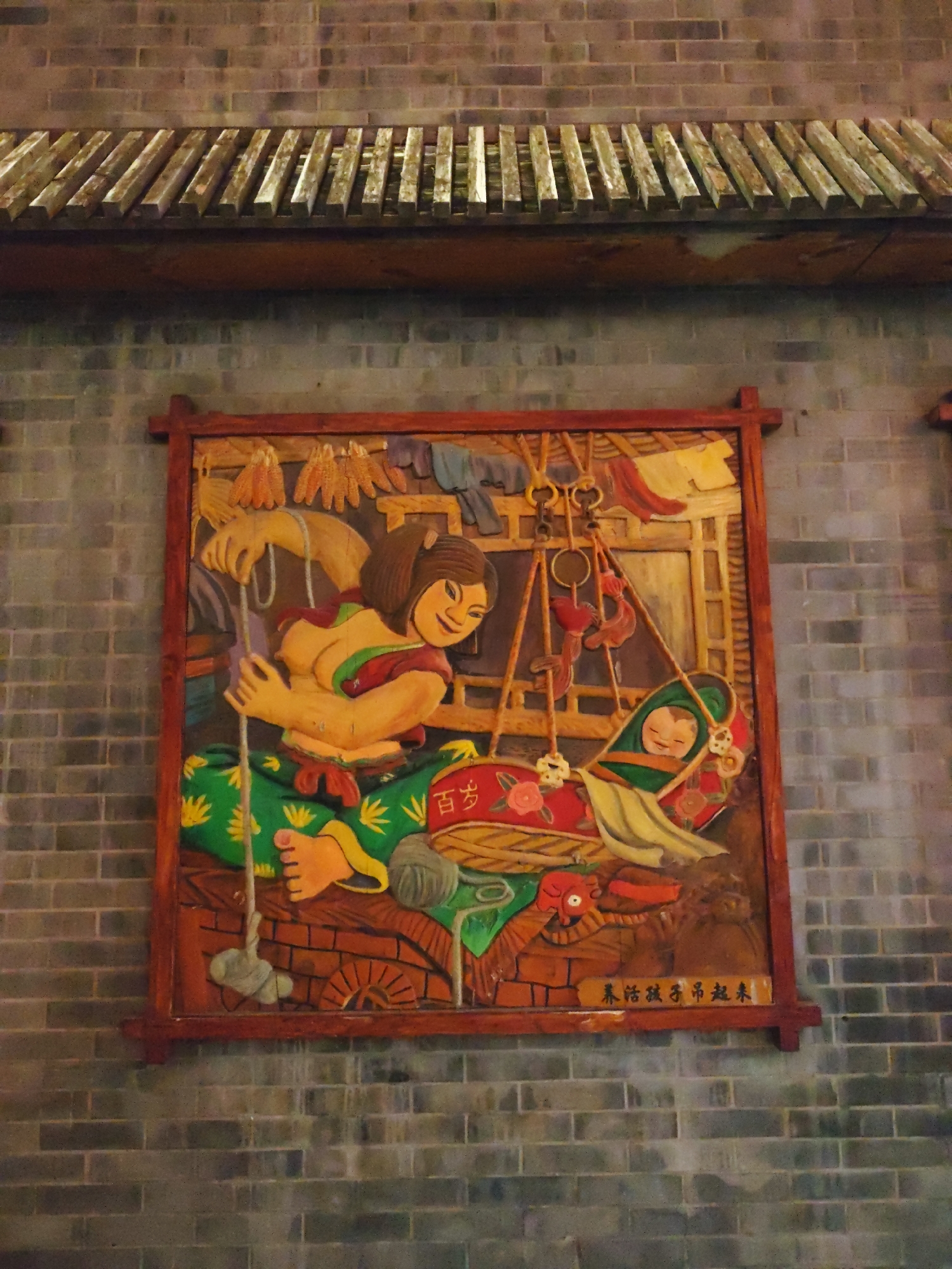 The image shows a colorful, carved wooden relief panel depicting a scene of domestic life. The panel is framed in dark wood and mounted on a gray brick wall, with a wooden eave structure visible above it.
The central figures in the relief are two people and a baby. On the left, a woman with dark hair and a smiling expression is actively engaged in rocking a cradle. She has her left arm raised, holding onto a rope that appears to be connected to the cradle. Her right arm is bent, with her hand near her chest. She is wearing a yellow top with red and green accents and green pants patterned with yellow flowers. Her feet are visible, with her toes pointing upwards.
To the right of the woman, a baby is comfortably nestled in a patterned cradle, which is suspended by ropes. The baby is smiling and appears to be wrapped in green and yellow fabric. The cradle itself is red and adorned with floral motifs.
The background of the relief suggests an indoor setting. Various objects are depicted, including what appear to be dried corn husks hanging from the top, various utensils and tools suspended on hooks, and clothing items like a yellow shirt and a red cloth hanging on a clothesline. There are also elements that suggest furniture or structural details, like wooden beams and brickwork.
At the bottom of the relief, there is a horizontal wooden bar with Chinese characters inscribed on it. These characters are not fully legible in the provided image, but they appear to be some form of inscription or title.
The overall style of the carving is somewhat stylized and folkloric, with bold colors and a focus on conveying a narrative of daily life and care. The brick wall and the wooden eave suggest the setting is likely an exterior wall of a traditional building, possibly in a rural or historical context within Lindian, China.