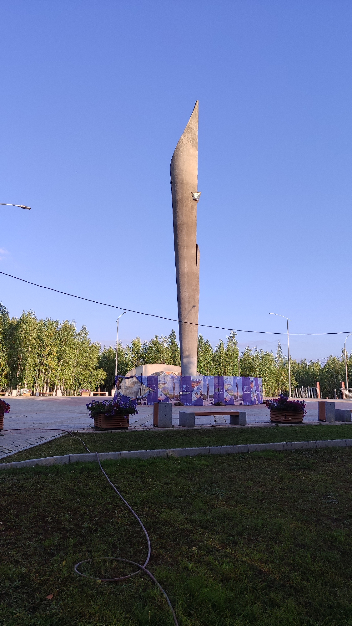 The image shows a tall, slender concrete monument with a sharp, angular top, set against a clear blue sky. The monument is located in Mirny, Russia, in an open, paved area surrounded by a line of deciduous trees. In front of the monument, a large banner with purple and blue graphics, featuring portraits and the number "70," is stretched between poles. Several benches are placed in front of the banner. To the left and right of the monument, there are large wooden planters filled with purple flowers. A dark electrical wire runs horizontally across the upper portion of the image. In the foreground, there is a patch of grass with a dark-colored hose coiled on it. The scene appears to be a public space, possibly a park or square, likely commemorating an event or historical figures. There are no people visible in the image.