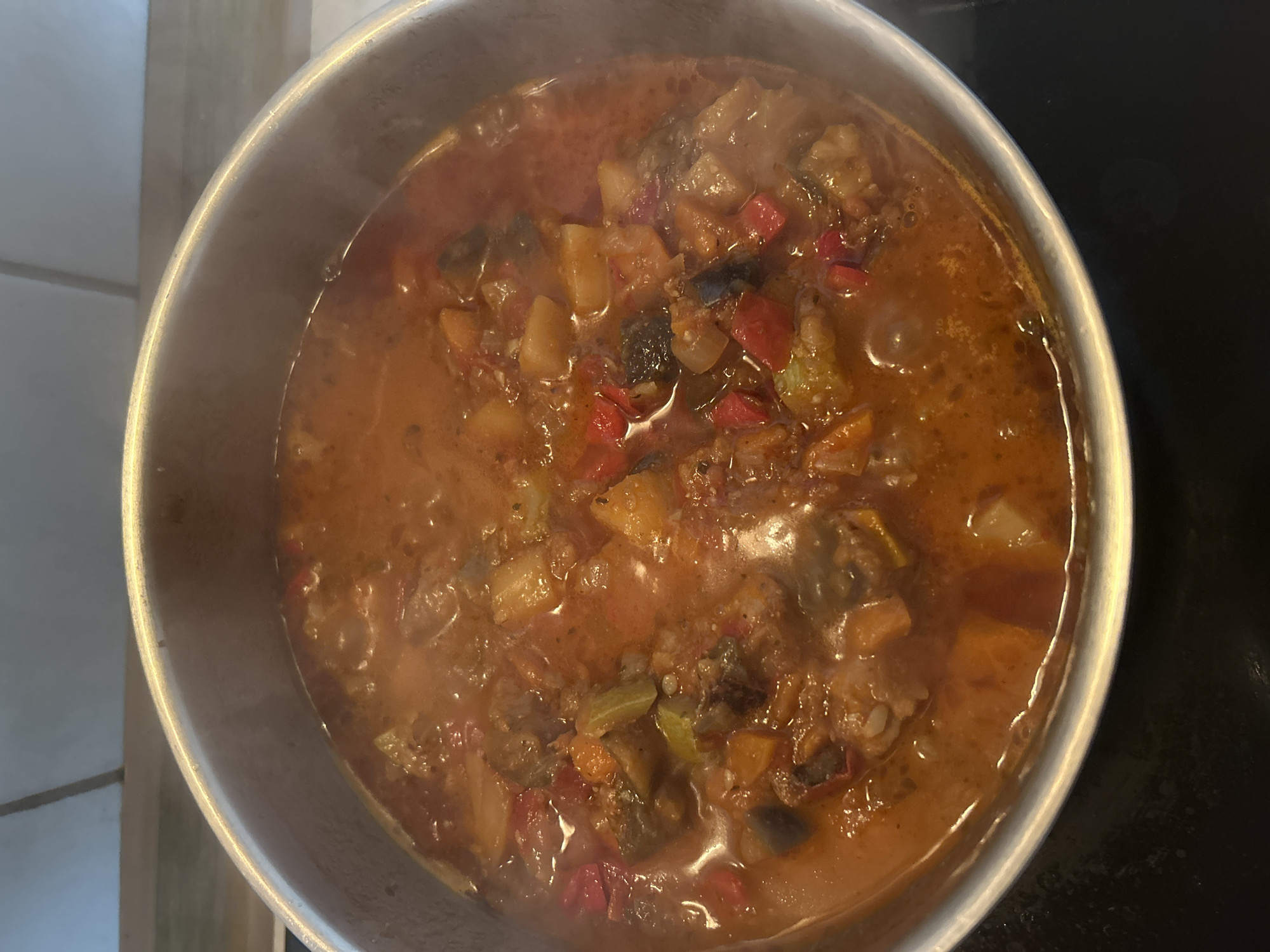 A top-down, medium shot captures a pot of simmering stew filled with diced vegetables including eggplant, bell peppers, and possibly zucchini, all in a rich, reddish-brown broth. The stew is steaming, indicating it is hot and actively cooking. The pot is a silver-colored metal, and it sits on a black stovetop. The background is slightly out of focus, showing a light-colored tiled surface and what appears to be a wall or cabinet. The scene is indoors and appears to be a kitchen environment. There is no indication of the city of Detmold, Germany, or any specific location within it. The image focuses solely on the cooking stew.