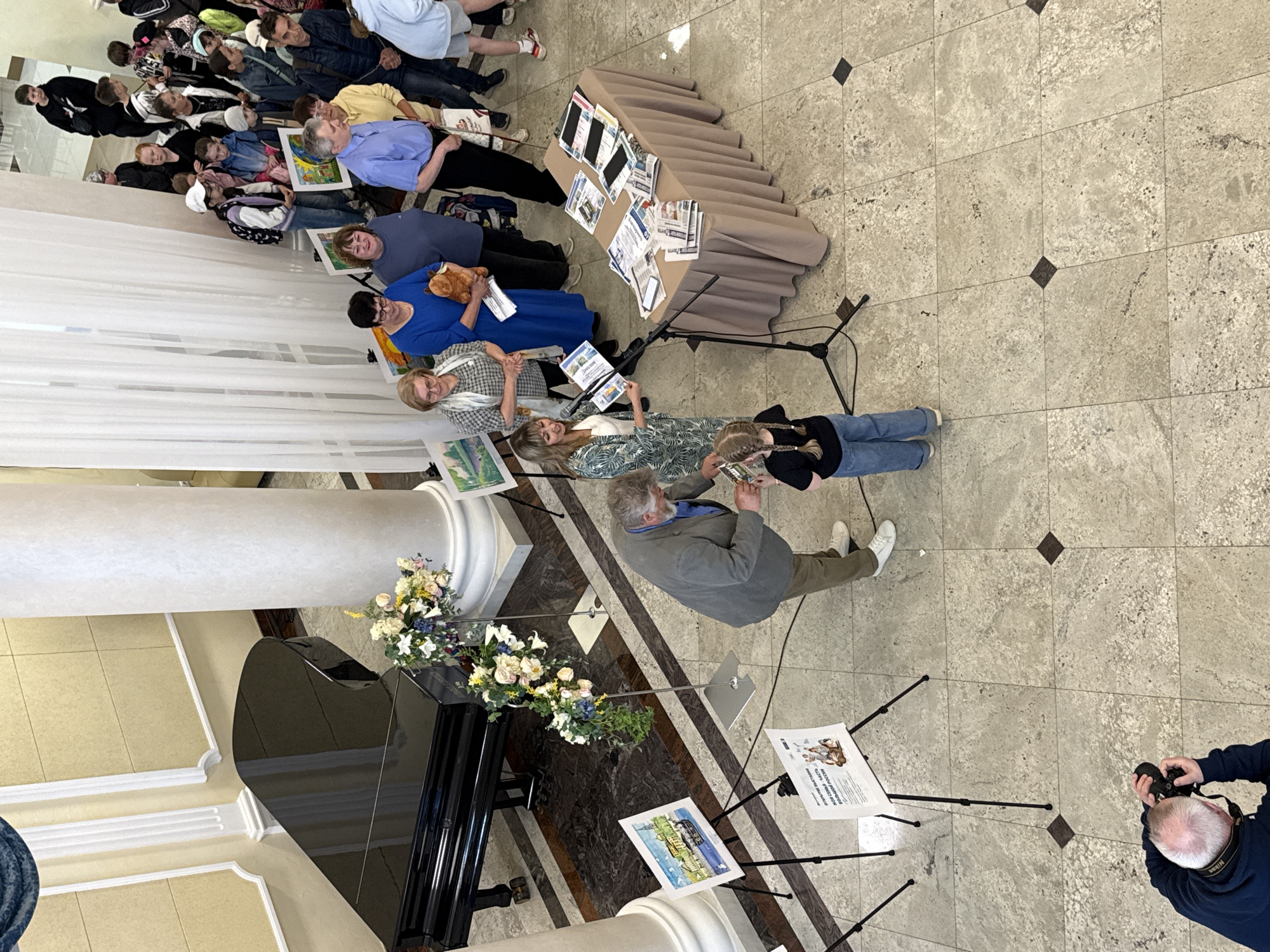 A high-angle shot shows a group of people inside what appears to be a gallery or public space, likely in Votkinsk, Russia, judging by the presence of promotional materials in the background. The floor is light-colored tile with dark accents. A black grand piano sits to the left, adorned with flower arrangements. A white column stands adjacent to the piano.
A crowd of people sits in rows, facing the camera, suggesting they are part of an audience. Some individuals hold artwork or objects. A table covered with a tan cloth displays papers and electronic devices, potentially tablets or phones. Microphones are positioned near the table, implying a presentation or event.
To the right, a man stands looking at a displayed artwork mounted on a tripod. Another man, likely a photographer, kneels and takes pictures with a camera. Tripods are used to display other pieces of artwork.