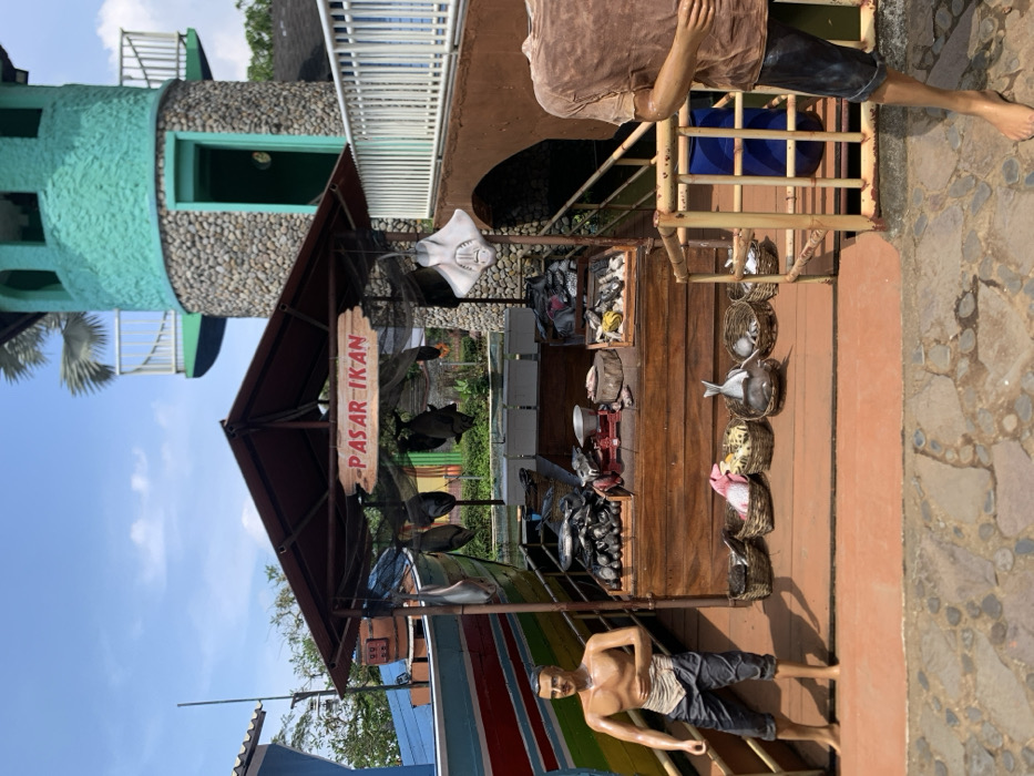 The image shows a fish market stall in the city of Batu, Indonesia, located at latitude -7.883 and longitude 112.526. The stall is made of wood and has a roof. It is located next to a small, wooden boat. The stall is decorated with a sign that says "PASAR IKAN" in red letters on a white background. It has a variety of fish on display, including stingrays, catfish, and tuna. There are baskets of fish on the counter and some hanging from the roof. A man in a blue t-shirt stands next to a railing. The stall is located next to a green building with a stone facade and a wooden railing. There is a blue barrel nearby. The ground is paved with cobbles and the sky is clear.