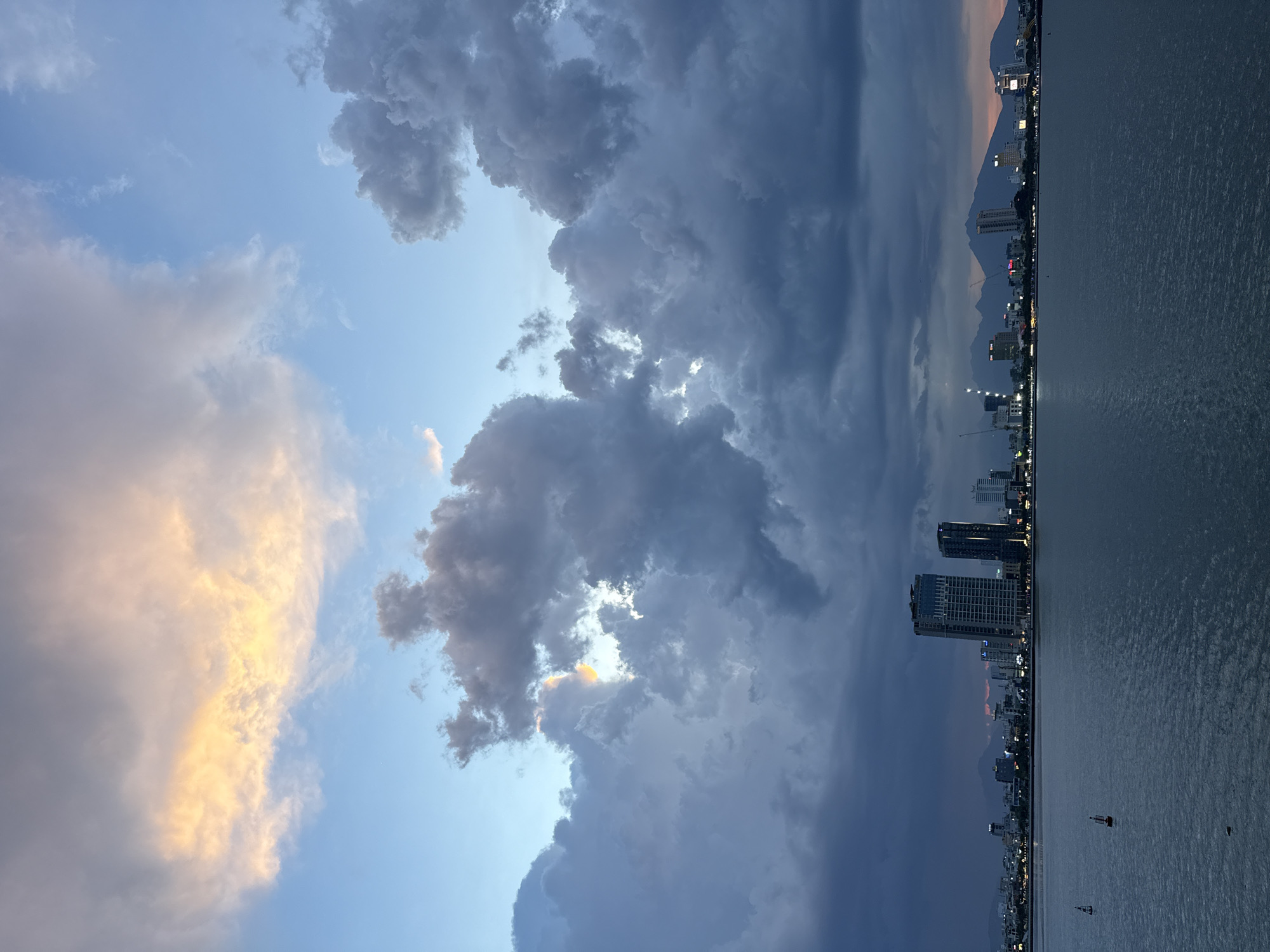 The image captures a dramatic twilight sky over the city of Đà Nẵng, Vietnam. Dominating the frame are a variety of clouds, some illuminated with a warm, golden hue from the setting sun, while others are dark and foreboding, suggesting an approaching storm. The sky transitions from a pale blue near the horizon to deeper shades of gray and indigo as the clouds thicken.
Below the sky, the Đà Nẵng coastline stretches out. The calm water of the sea or a large river reflects the muted light. Along the shore, a cityscape is visible, with numerous buildings of varying heights, some appearing as silhouetted shapes against the dusky sky, while others are beginning to show illuminated windows and lights, indicating the evening is settling in. In the distance, mountains form a soft, hazy backdrop. A few small boats or buoys are scattered on the water's surface.
The overall mood is one of transition and impending change, with the beauty of the sunset colors juxtaposed against the dark, heavy clouds. The time of day is clearly twilight, as suggested by the mix of natural light and artificial illumination. The location is identified as Đà Nẵng, Vietnam, and the presence of coastal scenery and urban development supports this. No visible text is present in the image.