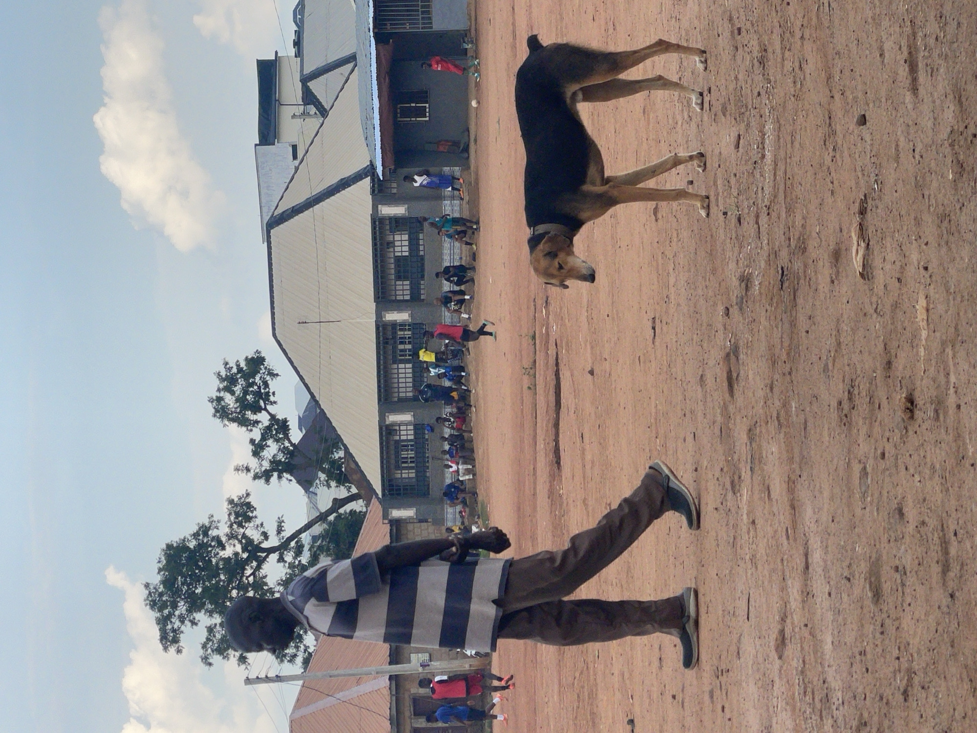 In the city of Kurunduma, Nigeria, a man in a striped shirt walks across a dirt field. To his right, a brown dog with black markings stands on the same field. In the background, various people are scattered around a building with a corrugated metal roof. Some people appear to be playing soccer, as evidenced by a ball visible on the ground. The environment is dusty and sparsely vegetated, with some buildings and trees visible on the horizon. The sky is partly cloudy.