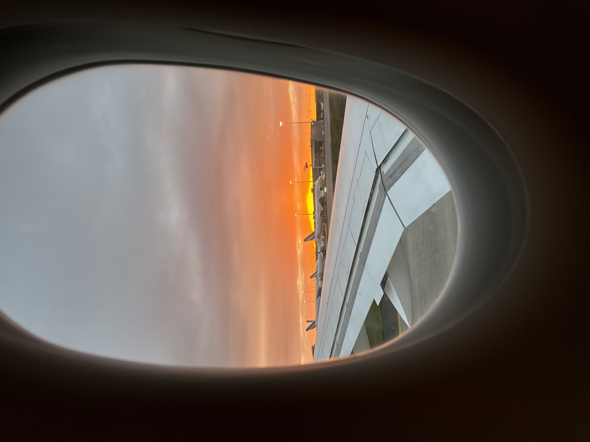 The image shows a view from an airplane window. Visible through the window is a vibrant orange sky, likely during sunrise or sunset. An airplane wing is also visible on the right side of the window. Beyond the wing, the ground contains airport infrastructure, including buildings and lampposts. Based on location, the scene likely captures an image taken while on a plane at Charles De Gaulle airport located near Le Mesnil-Amelot, France.