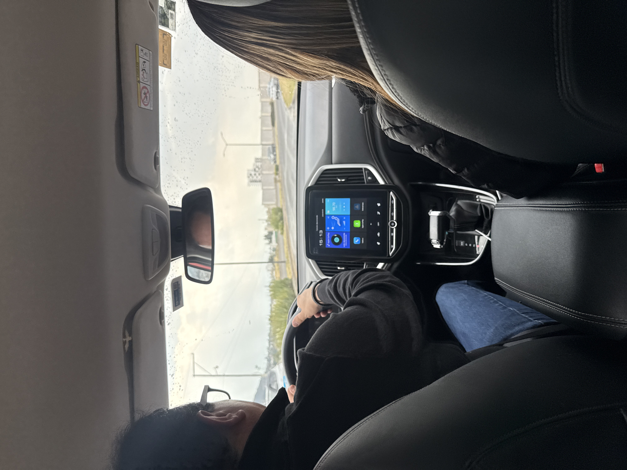 The image depicts an interior view of a vehicle, likely a car, in Santa Clara Ocoyucan, Mexico. The perspective is from the rear passenger seat, looking towards the front. The driver, wearing glasses and a dark-colored jacket, is gripping the steering wheel. The driver’s face is partially visible. The passenger seat is occupied by an individual with long hair, wearing a dark jacket, with only the back of their head visible. The dashboard features a digital display showing 15:13. The exterior is visible through the windshield, revealing an overcast day with a road and sparse vegetation. The vehicle’s interior is characterized by black leather seating and dashboard elements. The driver appears to be in control of the car, though the precise actions beyond steering are unclear. The environment appears to be urban outskirts with limited traffic.
