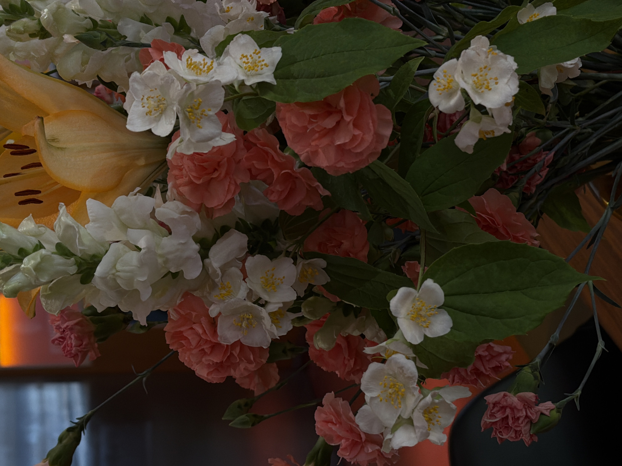 The image is a close-up of a floral arrangement featuring a variety of flowers. Predominant are light peach-colored carnations and white jasmine flowers with yellow stamens. There are also white snapdragons and a partially visible orange lily on the left. Green leaves are interspersed throughout the arrangement. The background is dark and blurred, suggesting an indoor setting, with faint hints of warm light. No people are visible. The location is not evident from the image and cannot be determined as Yerevan, Armenia.