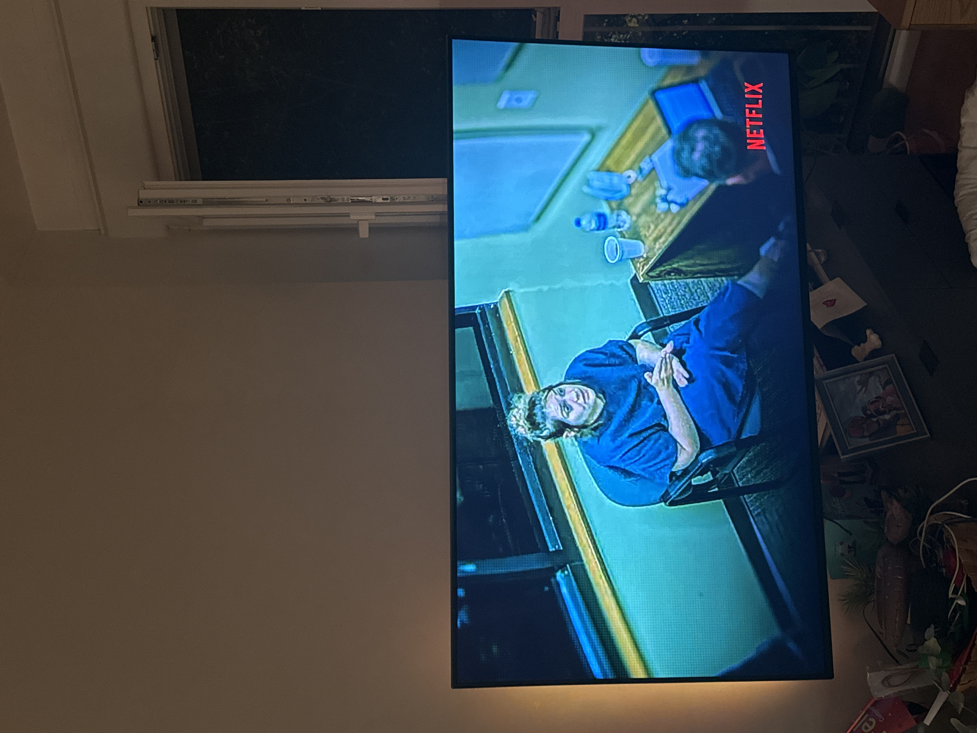 The image shows a television screen displaying a scene from a Netflix program. On the screen, a woman in a blue jumpsuit is seated in a chair in what appears to be an interrogation room. She has her hair pulled up in a bun and is looking to her left with a concerned expression. A table is in front of her with a plastic cup, a bottle of water, and a laptop. Another person's head is visible to the right of the table, facing the woman. The Netflix logo is displayed in red on the lower right side of the screen. The television is placed in a dimly lit room, with a window visible in the background, showing darkness outside. Below the television, there are various objects on a surface, including framed pictures, a basket, and some greenery. There is no information available about the location being the city of Murcia, Spain.