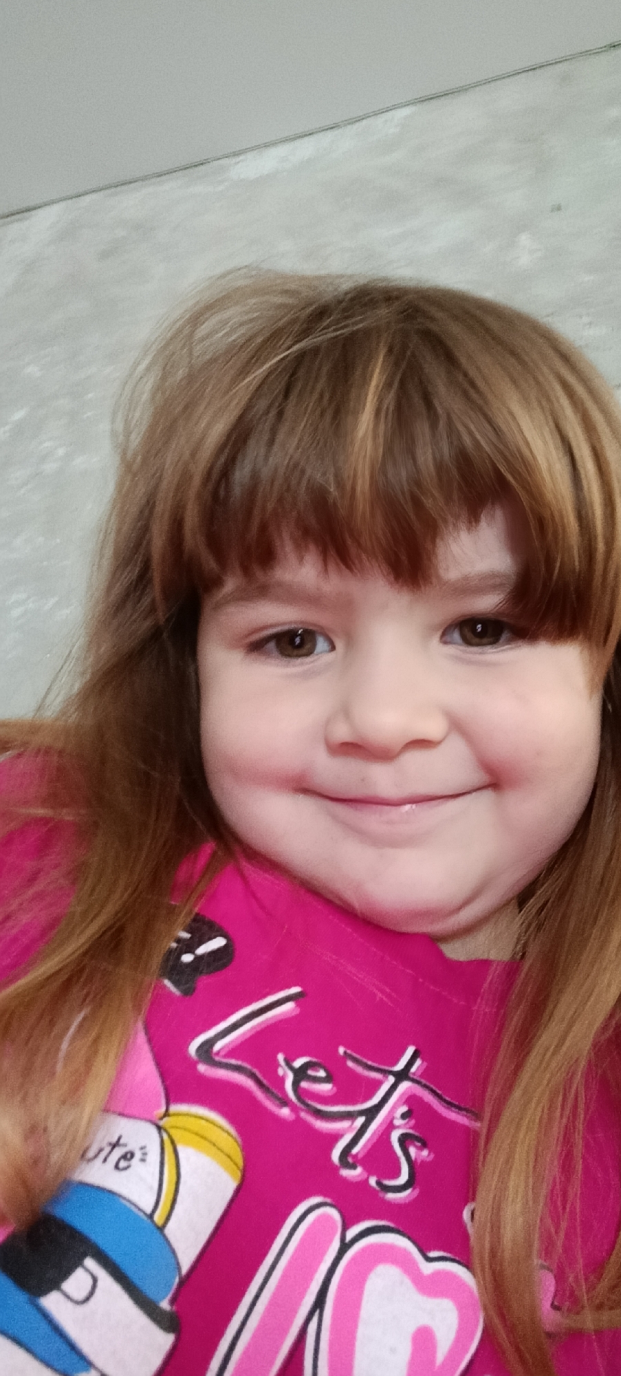 A young girl with brown hair and brown eyes is captured in a close-up, selfie-style portrait. She is wearing a bright pink t-shirt with a graphic design. The t-shirt features the word "Let's" in a stylized font, along with other cartoon-like graphics. Her hair is long and appears to be styled with bangs. The background is a neutral, textured gray wall. The lighting is soft and even, illuminating her face. The image is taken from a slightly upward angle. There is no indication of the specific location within Хохлы, Russia.