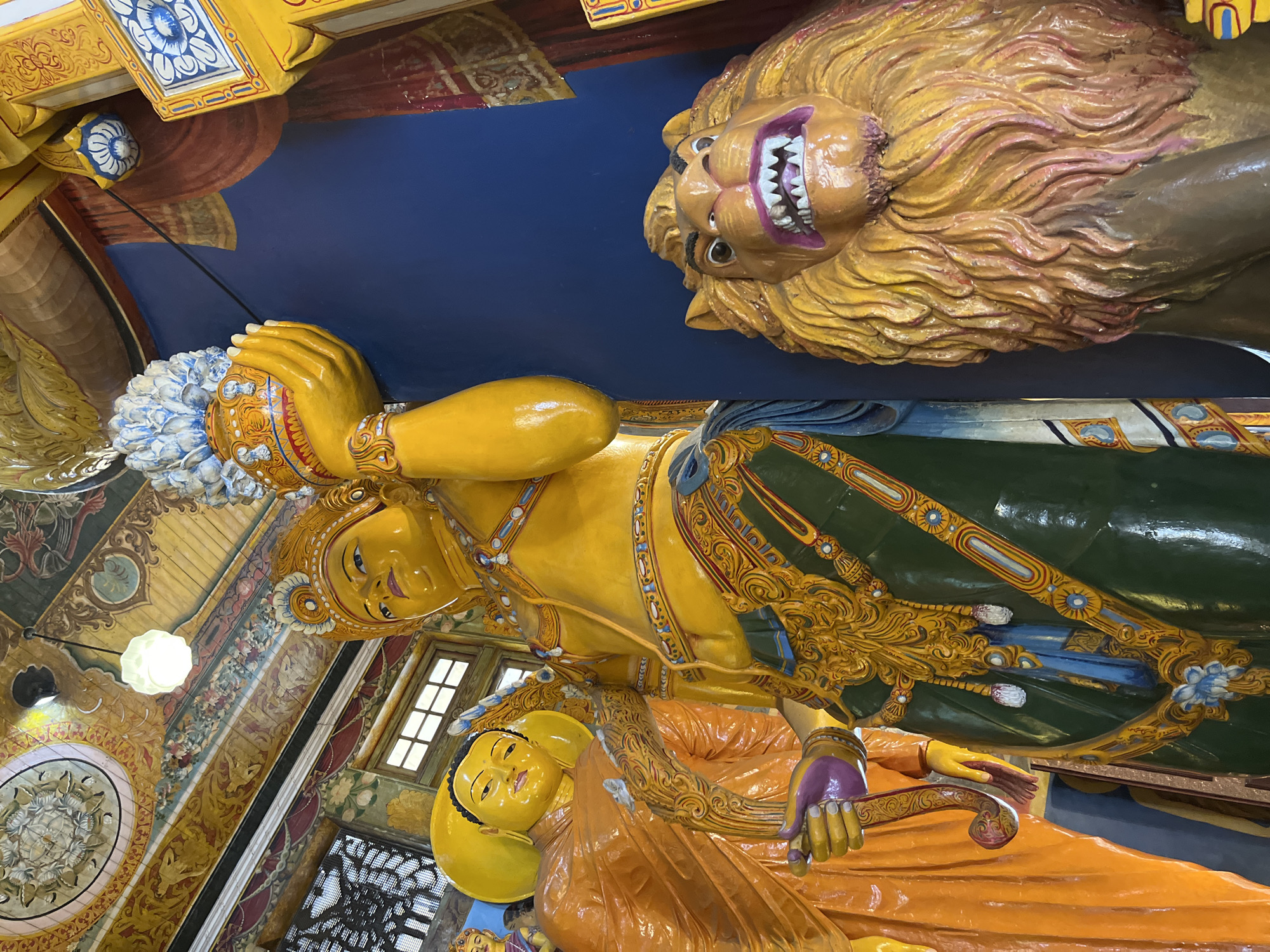 The image captures a close-up, low-angle view of vibrant statues within a Buddhist temple in Colombo, Sri Lanka. In the foreground to the right, a striking, fierce-looking lion statue with a golden mane and bared teeth is prominently displayed against a dark blue wall. To the left and slightly behind the lion, a tall, golden statue of a figure, possibly a deity or a significant Buddhist figure, stands adorned in a green and gold garment, holding a golden urn with a blue and white decorative top in its right hand. To the far left and further back, another large statue of a serene figure, likely a Buddha, is visible, wearing an orange robe. The background reveals ornate, colorful ceiling decorations with intricate patterns and motifs, and parts of windows with latticework. The overall scene is rich in color and detail, indicative of traditional temple artistry.