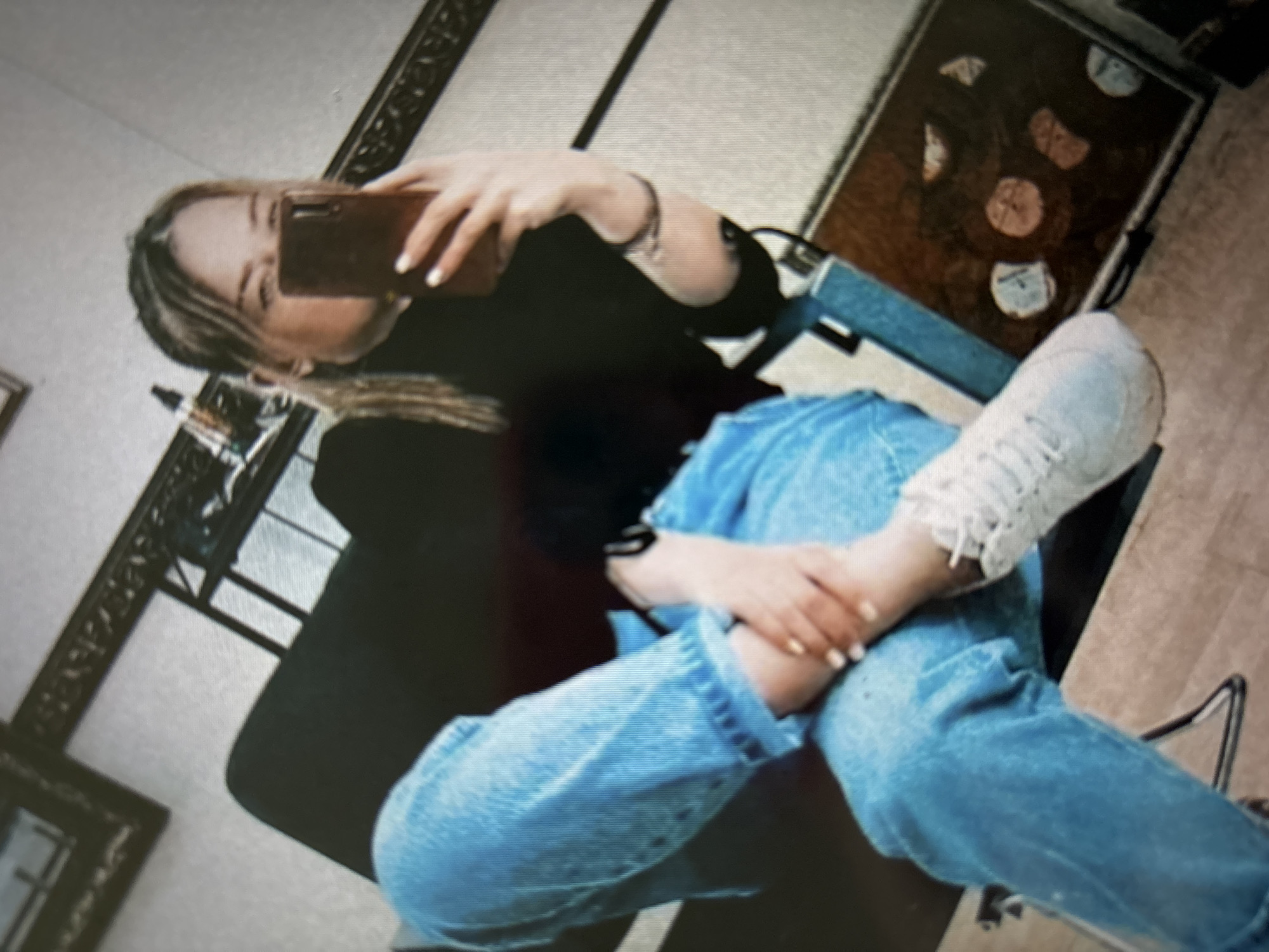 In a medium shot, a young woman with blonde hair pulled into a ponytail is seated indoors, taking a selfie with a smartphone held in her right hand. She is wearing a black turtleneck and light-wash denim jeans. Her left leg is crossed over her right, and her left foot is resting on the floor, clad in a white sneaker. A dark-colored object, possibly a chair or bench, is behind her, and a framed picture is visible on the wall in the upper left corner. A dark metal railing runs horizontally in the background, and another framed item, appearing to be a collage of circular objects, is visible on the floor to the right. The lighting in the room is dim. The specific location within Novosibirsk, Russia, cannot be determined from the image.