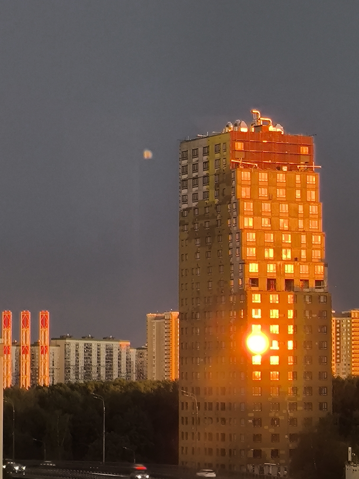 The image captures a cityscape at dusk, characterized by a muted, overcast sky. A prominent, unfinished high-rise building dominates the right side of the frame, its facade a mixture of construction materials and yellowing insulation. The setting sun casts a brilliant orange glow upon the building, illuminating its numerous windows and creating a striking reflection that appears as a bright, circular orb in the middle section of the structure. The rooftop of this building features exposed piping and ventilation systems, further emphasizing its incomplete state.
To the left of the main building, a series of other residential high-rises are visible, their windows appearing as small, dark rectangles against the subdued sky. Three tall, slender structures with red and orange illuminated panels stand in the mid-left background, adding a vertical accent to the urban landscape. A dense canopy of dark green trees forms a natural barrier behind these buildings, obscuring the lower portions of the structures and the ground.
In the foreground, a highway or elevated road is partially visible, with a few blurred vehicles indicating movement. Streetlights are also present, casting a dim light on the road. The overall impression is one of an urban environment transitioning from day to night, with the dramatic lighting on the construction site being the most notable element. The presence of construction materials and the unfinished appearance of the main building suggest an active development in this part of the city.