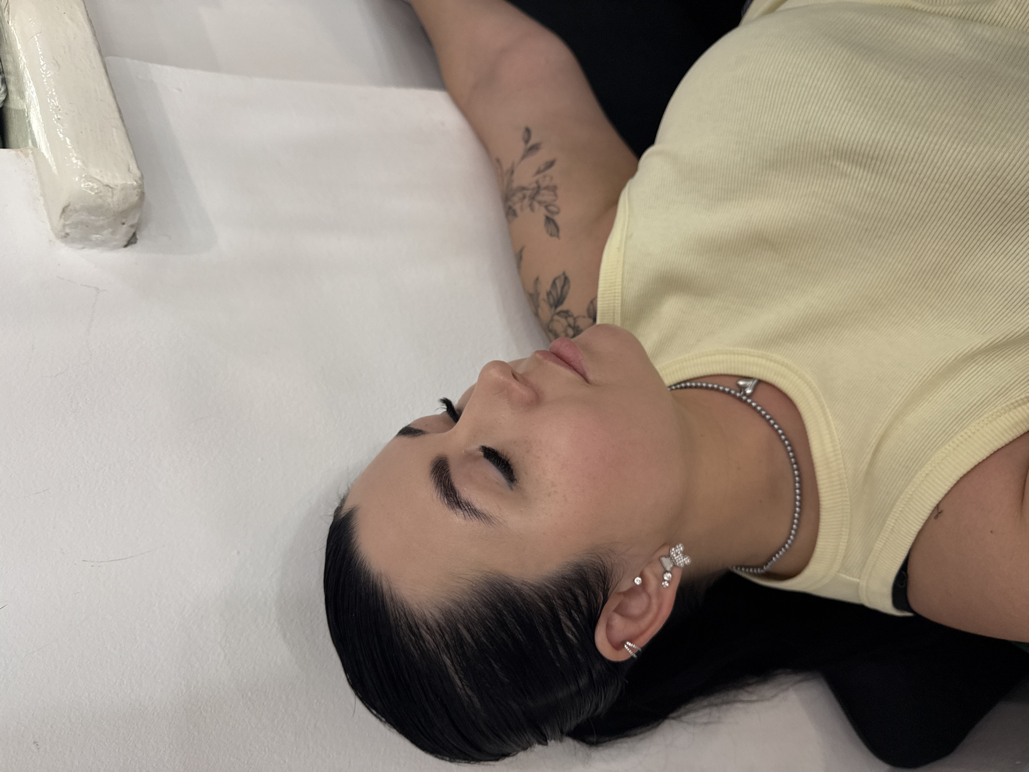 A woman with dark hair, slicked back, is lying on a white surface, possibly a bed or table. She is wearing a yellow ribbed tank top and a silver beaded necklace. She has multiple piercings in her left ear and a tattoo on her left arm. The location in the city of Chaikovsky, Russia, is not evident in this image.