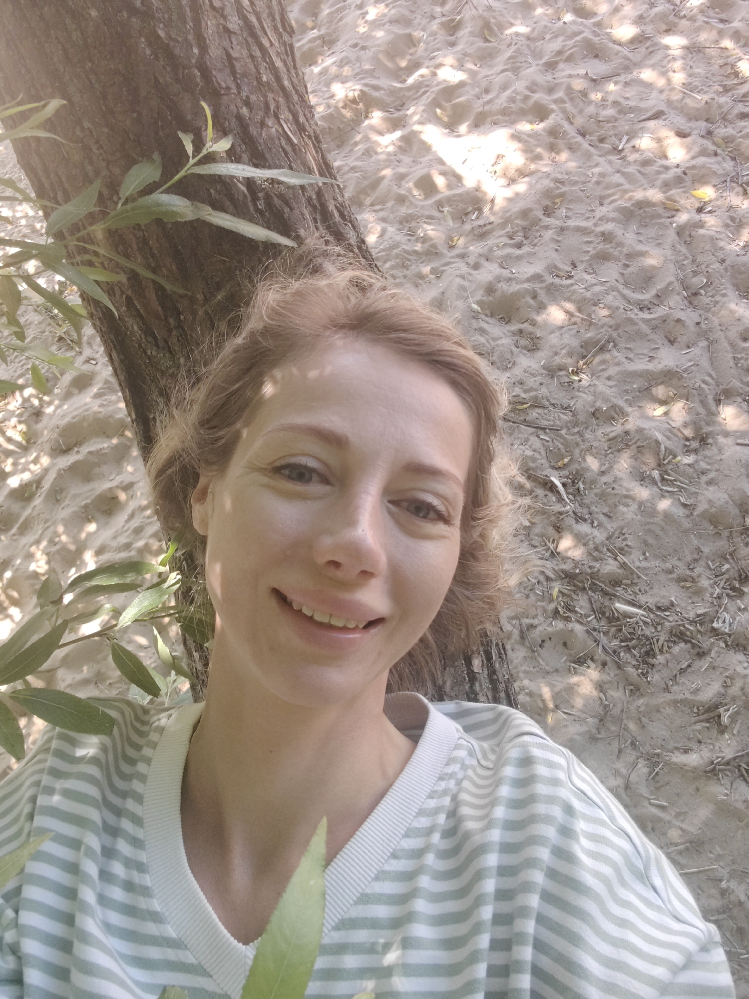 An outdoor, eye-level, medium shot shows a young adult Caucasian woman with light brown hair, fair skin, and blue eyes, smiling. She is wearing a light green and white striped sweatshirt. She is positioned leaning against a tree trunk, with her right side slightly towards the camera. The background consists of sandy ground with scattered dry leaves and twigs, and dappled sunlight shining through unseen foliage. The overall environment appears to be a park or natural area. The location is not specified to be Saint Petersburg, Russia.