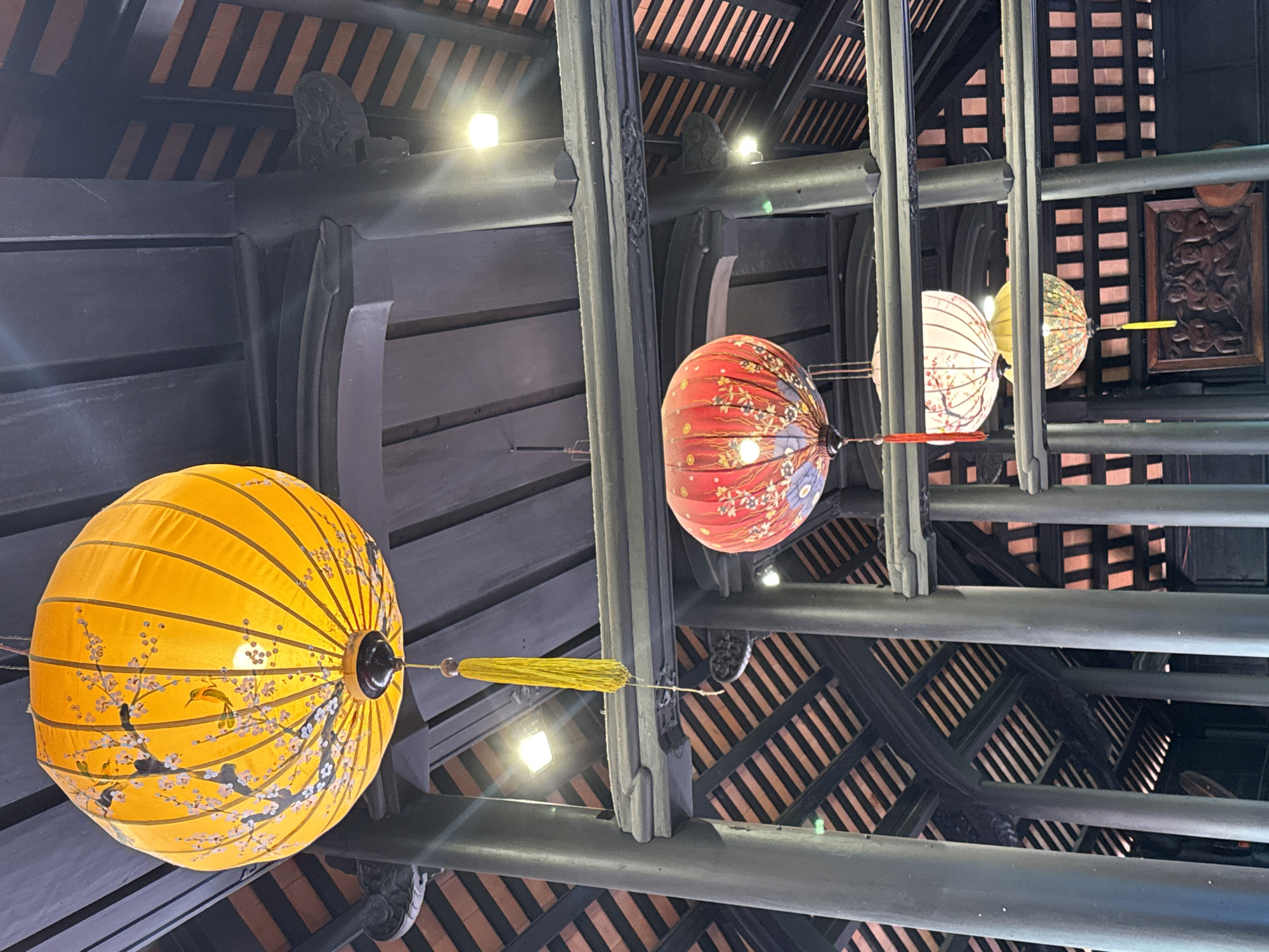 The image shows a view looking upward at a ceiling with traditional Vietnamese lanterns hanging from it. The location is possibly in the city of Bac Nha Trang Ward, Vietnam. The ceiling is constructed of dark wooden beams arranged in a grid pattern, with narrow gaps between the slats. Four lanterns are visible, each a different color (yellow, red, and two white). The lanterns are round and decorated with floral or abstract designs. Each lantern has a decorative tassel or string hanging from it. The beams above are intricately carved.