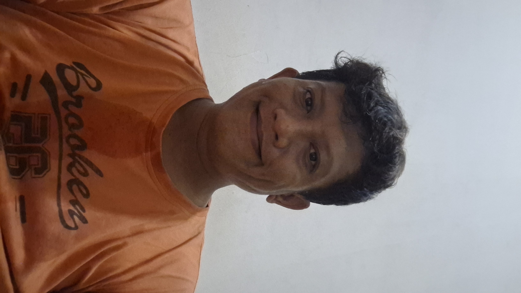 A medium shot shows a man in an orange t-shirt looking directly at the camera against a plain, light-colored background. The man has dark, wavy hair and a light brown complexion. He is smiling, with visible dimples. The t-shirt has a dark graphic design on the chest that appears to say "Brooken 26". The image appears to have been taken indoors, likely in a residential setting in Cibinong Hilir, Indonesia, given the man's appearance and the simple backdrop.