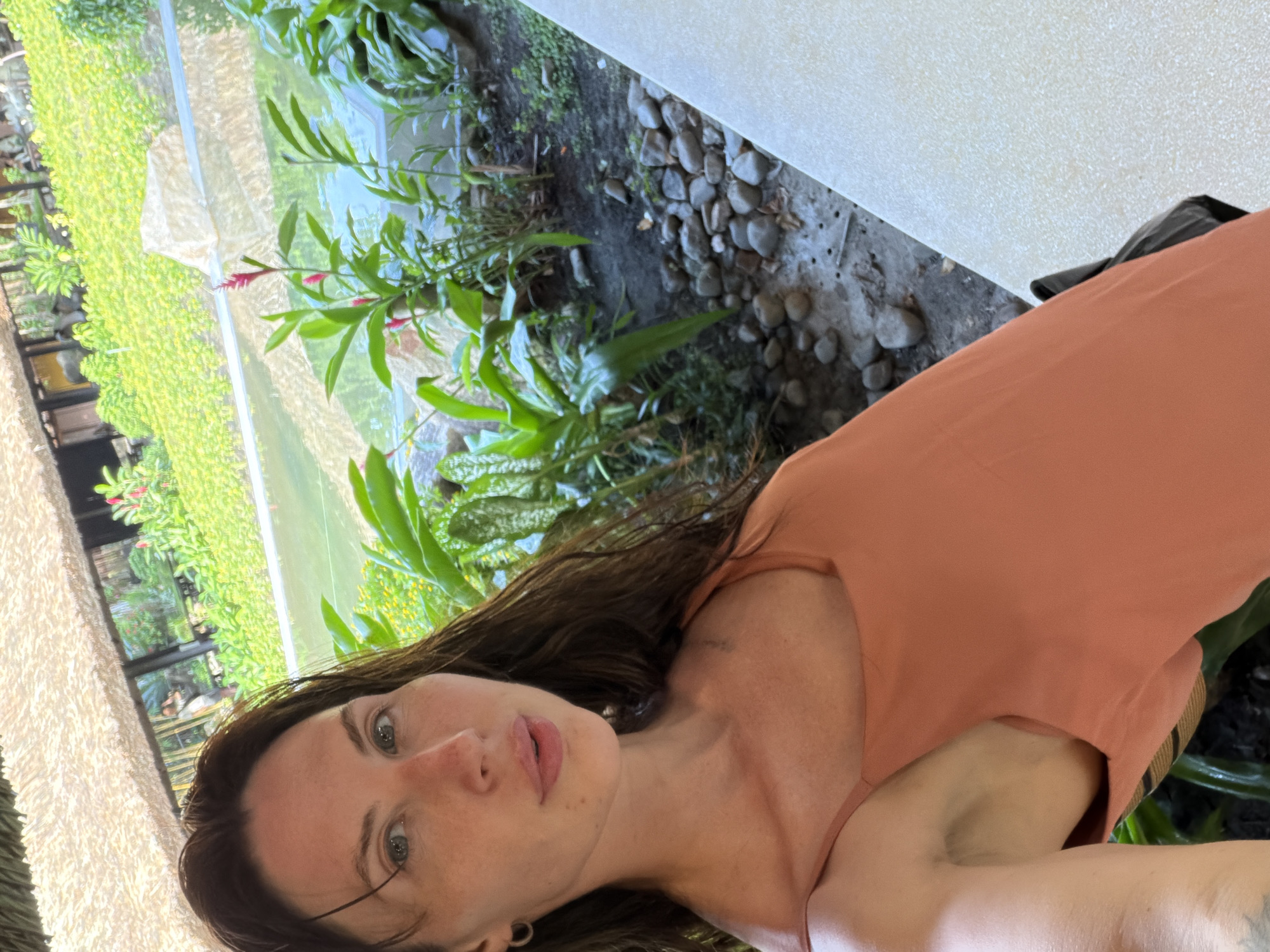 A woman with long brown hair and light skin is looking at the camera. She is wearing a peach-colored tank top. The background shows a lush green garden with a body of water. The garden appears to be outdoors, possibly at a resort or spa, given the tropical foliage and the thatched roof structure visible in the distance. The overall scene suggests a relaxed, vacation-like atmosphere. There is no specific indication of the city of Tay Nha Trang Ward, Vietnam, in the image.