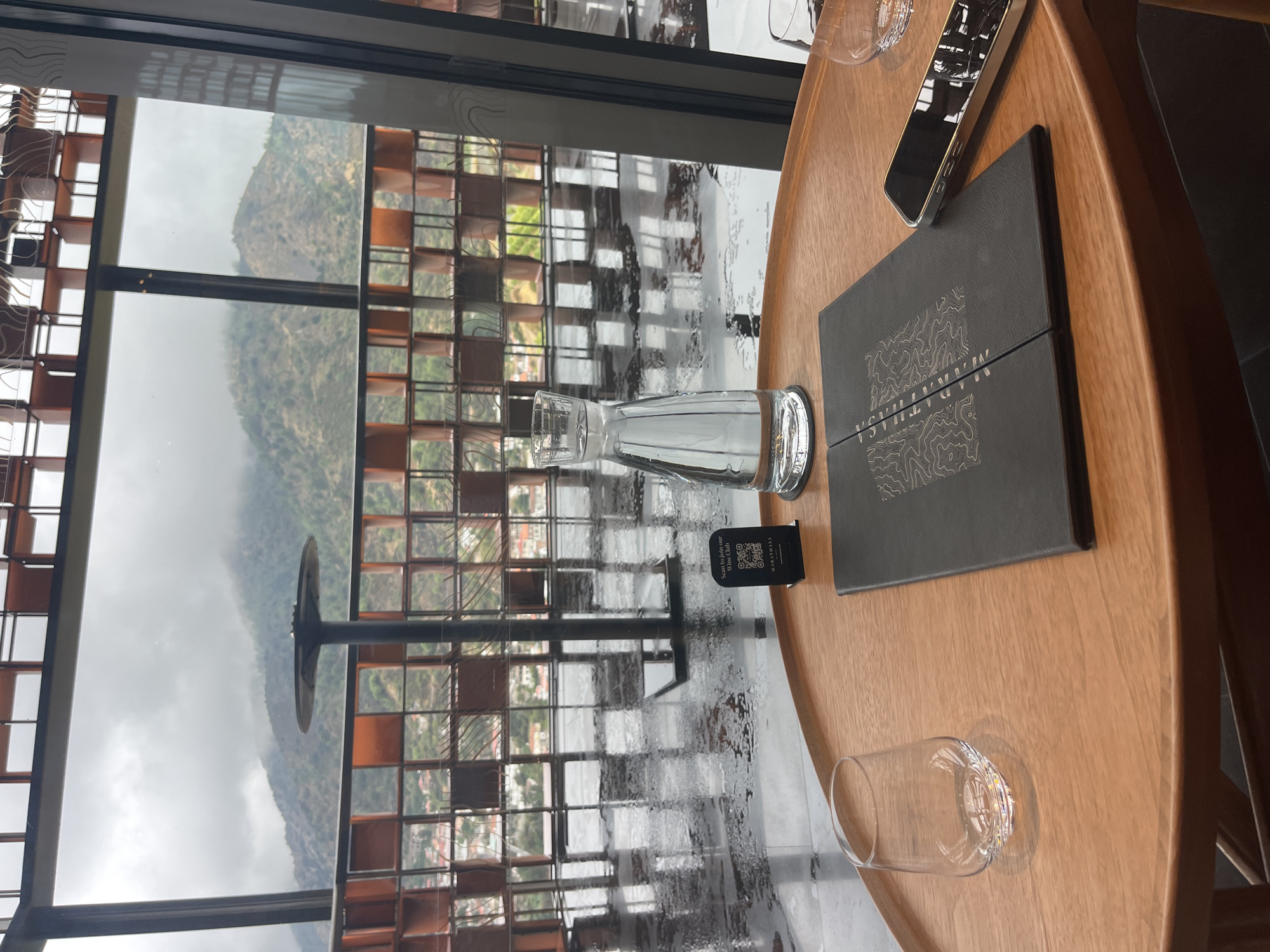 A close-up, eye-level, medium shot captures a wooden table in a restaurant with a view of the Troodos Mountains in Kalopanagiotis, Cyprus. The table, set for a meal, holds a clear glass, a closed menu with a topographical map design, a smartphone, and a carafe filled with water. The floor appears wet and reflective, possibly from recent rain. Beyond the table, large windows reveal a panoramic vista of lush green mountains shrouded in mist, with a town nestled in the valley below. A decorative architectural element, possibly a wine rack or screen, with alternating clear and orange panels, frames the outdoor view. The overall mood is serene and contemplative, with the natural beauty of Cyprus serving as a striking backdrop to the intimate dining setting.
