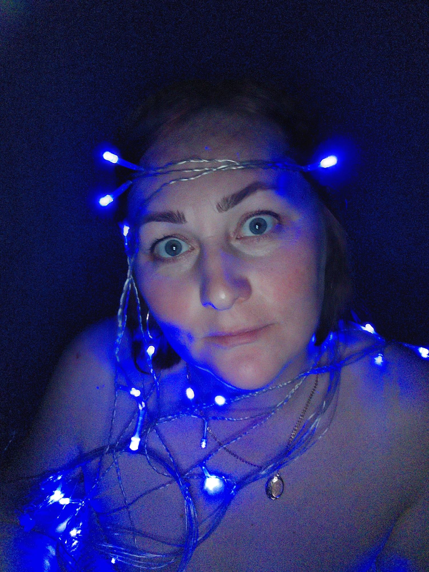 A medium-close-up shot shows a Caucasian woman with light skin, blue eyes, and light brown hair illuminated by strands of blue LED fairy lights. Some lights are wrapped around her head like a crown, and others are draped around her neck and shoulders. She wears a delicate necklace with a small, circular pendant. Her expression is one of mild surprise or bemusement, with raised eyebrows and a slight pursing of her lips. The background is dark, providing high contrast to the bright blue lights. The location is not discernible from the image.