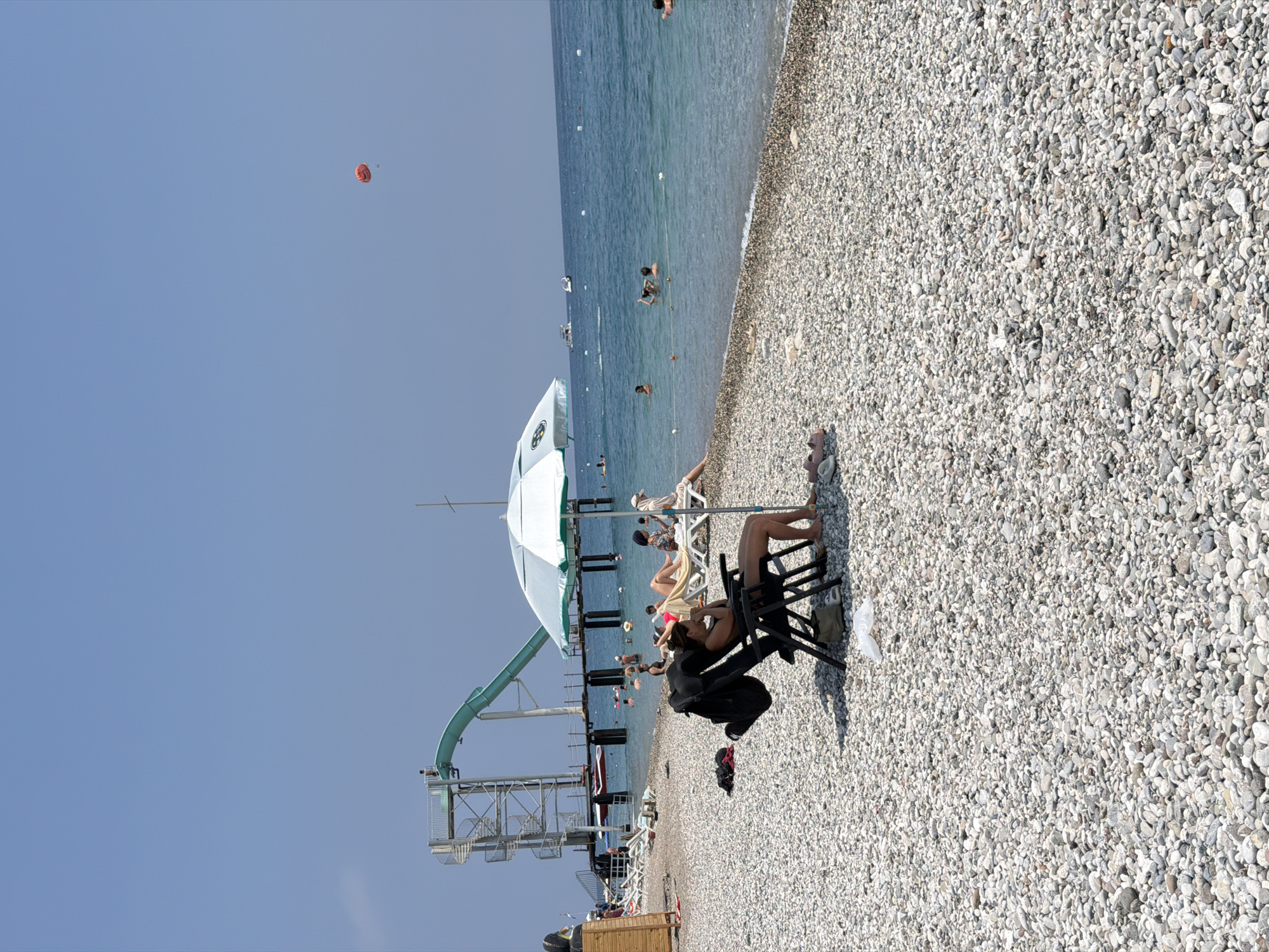 The image captures a sunny day at a pebble beach in Kemer, Turkey. In the foreground, a woman is seated in a black beach chair under a blue and white umbrella, appearing relaxed. Her belongings, including a black bag, are placed near the chair. The beach is covered with smooth, grey pebbles. In the midground, several people are enjoying the sea, swimming and wading. Further back, a large water slide structure with a spiral staircase rises above the water, connected to a pier. Boats are visible on the water, and a red parachute is descending from the clear blue sky in the upper right quadrant of the image. The sea is a vibrant blue, with gentle waves lapping the shore. The overall atmosphere is one of leisure and vacation activities.