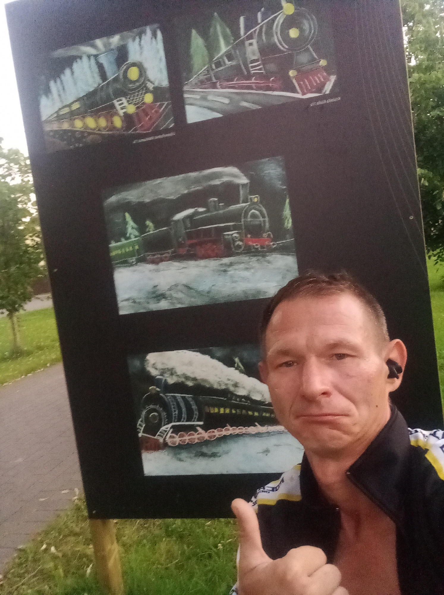 A man, wearing an open, unzipped jacket with a yellow and white stripe design and black earbuds, is taking a selfie in front of a large black sign displaying three paintings of steam locomotives. He is giving a thumbs-up gesture. The sign is outdoors, leaning against a wooden post, with grass and a paved path visible in the background. The paintings are in a colorful, somewhat stylized artistic depiction. The overall scene suggests a public display or outdoor art installation, possibly in a park or recreational area.