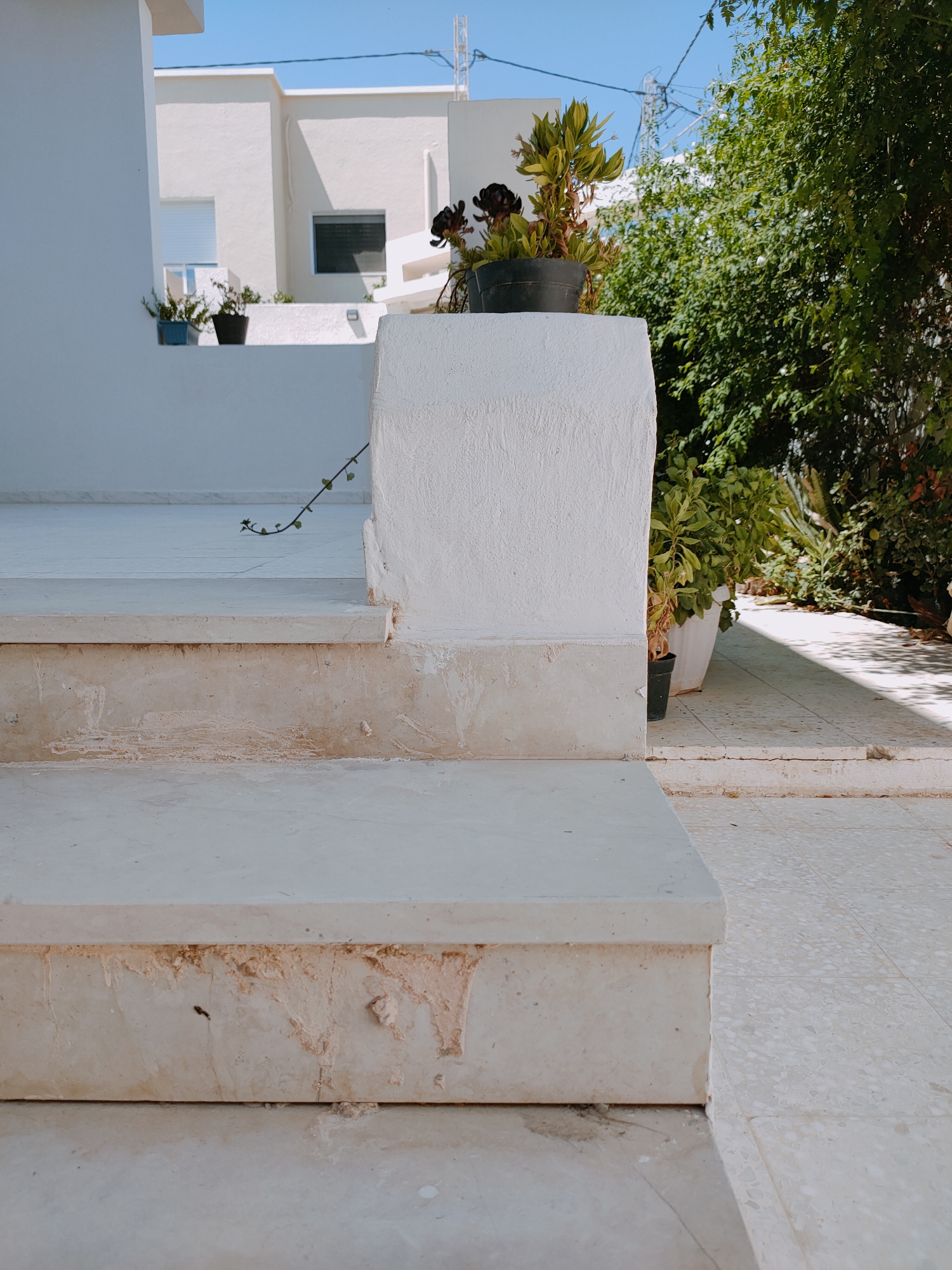 The image shows a close-up of an outdoor staircase made of light-colored stone, likely marble or travertine, in a sunny environment. The steps are wide and appear worn in places, with some evidence of staining or discoloration, particularly on the risers. The foreground is dominated by the staircase, with the first step filling the bottom of the frame.
Behind the staircase, a white, textured wall and a white railing are visible. To the right of the staircase, a paved area with small, mosaic-like tiles leads away into the background. Lush green foliage and trees create a natural backdrop, with dappled sunlight casting shadows on the ground. A few potted plants are strategically placed near the white structures, one containing a dark-leaved succulent.
In the distance, a modern white building with clean lines and geometric shapes is visible. It features windows, one with a roller blind and another with dark glass. The sky above is a clear, bright blue. Electrical wires and a utility pole are visible against the sky, suggesting an urban or suburban setting. The overall impression is one of a well-maintained, possibly residential, outdoor space in a Mediterranean or North African setting, consistent with the city of Ariana, Tunisia. There are no people or animals present in the image.