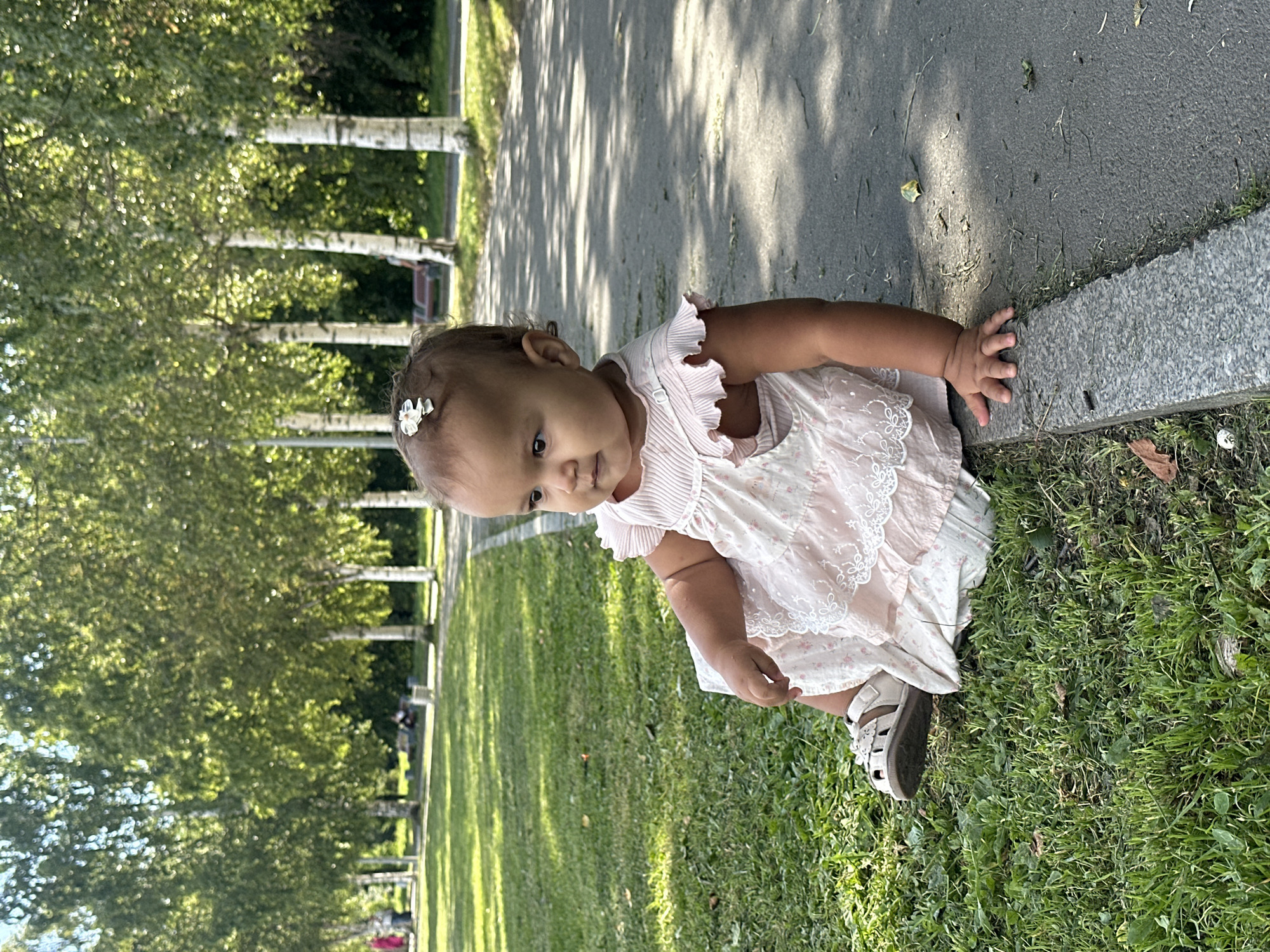 A young baby girl with a white flower clip in her hair is sitting on the grass next to a paved pathway in a park. She is wearing a light pink dress with lace trim and white sandals. The pathway is bordered by a granite curb. In the background, there are several birch trees with green leaves, and the sun is shining, casting shadows on the ground. The park appears to be in a city, with hints of buildings or structures in the far distance. The overall scene suggests a peaceful, sunny day in a recreational area. This park is likely the Onega Embankment in Petrozavodsk, Russia, as indicated by the characteristic birch trees and the layout of the waterfront park.