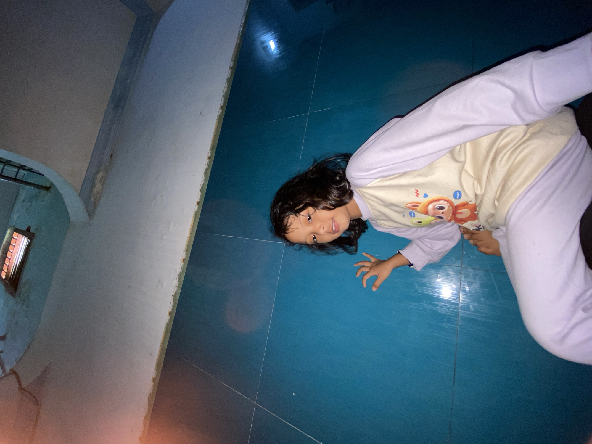 A young girl is seated indoors in Gelam, Indonesia, looking directly at the viewer with a slight smile. She has dark, wavy, shoulder-length hair and medium skin tone. She is wearing long-sleeved pajamas: a cream-colored top with light purple raglan sleeves and a graphic featuring an orange, rabbit-like cartoon character, a green frog, and a yellow chick, paired with solid light purple pants. Her bare feet are visible, and her right hand is raised, fingers slightly splayed.
The background consists primarily of a vibrant teal-colored tiled wall with visible white grout lines forming a grid pattern. Reflections and vertical streaks from an artificial light source are apparent on the tiled surface, particularly on the right side. Above the tiled wall, a light grey or off-white upper wall segment or ceiling transition shows some textural inconsistencies and discoloration near the border. The ceiling curves upwards, revealing a dark metal railing on the upper left, suggesting an adjacent staircase or upper level. In the upper right corner, a rectangular vent or small window with several horizontal orange-red slats is visible. An orange lens flare is present in the upper-left quadrant of the image.