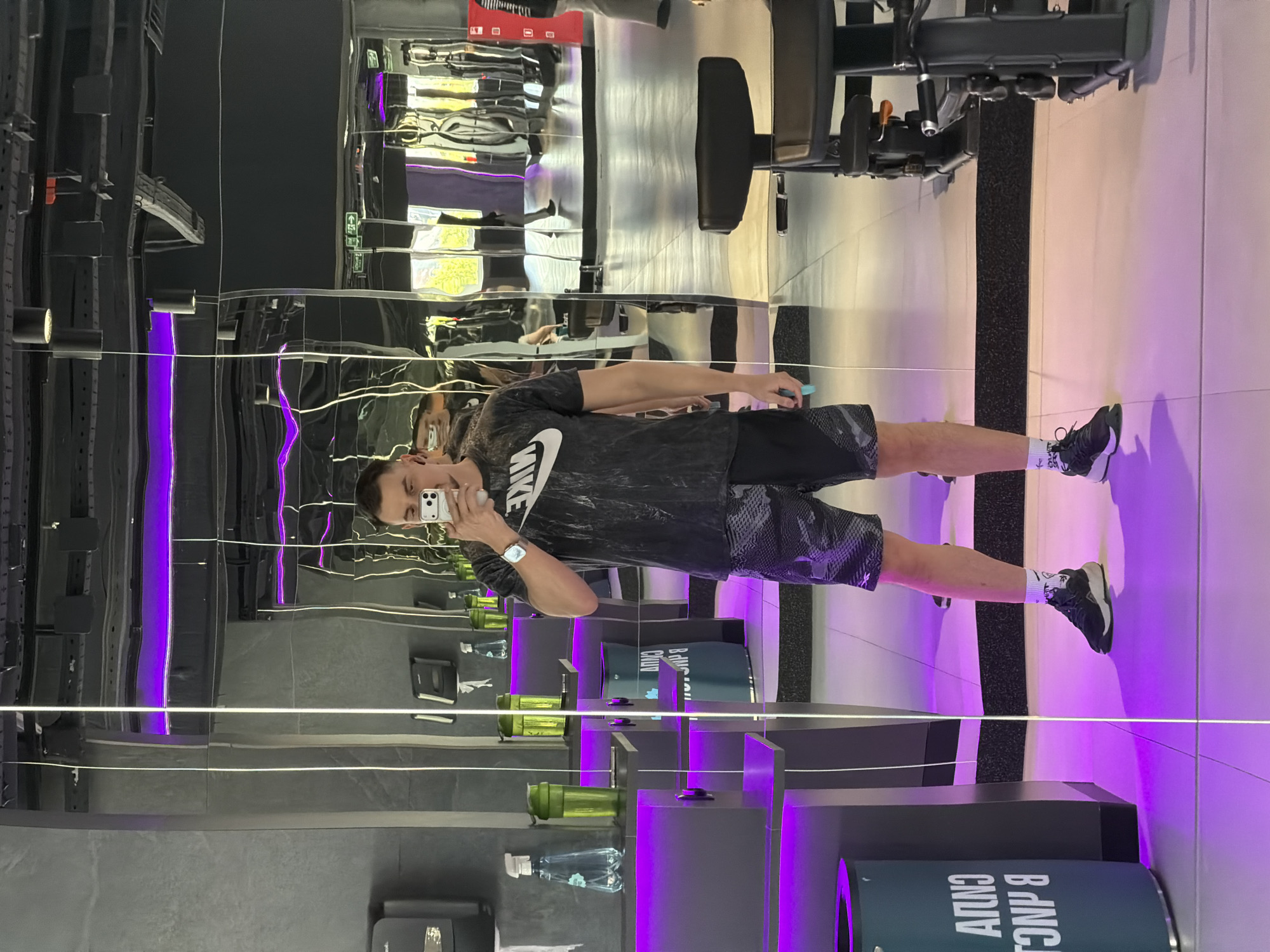 A man stands in a gym with mirrored walls and purple lighting. He is wearing a dark-colored Nike t-shirt, black shorts with a geometric pattern, and athletic shoes with white socks. He is holding a smartphone and taking a selfie in the mirror. Various water bottles, containers, and other gym-related items are visible on shelves and in the background. The gym appears modern with its gray tones and neon lighting. The gym is located in Krasnoyarsk, Russia.