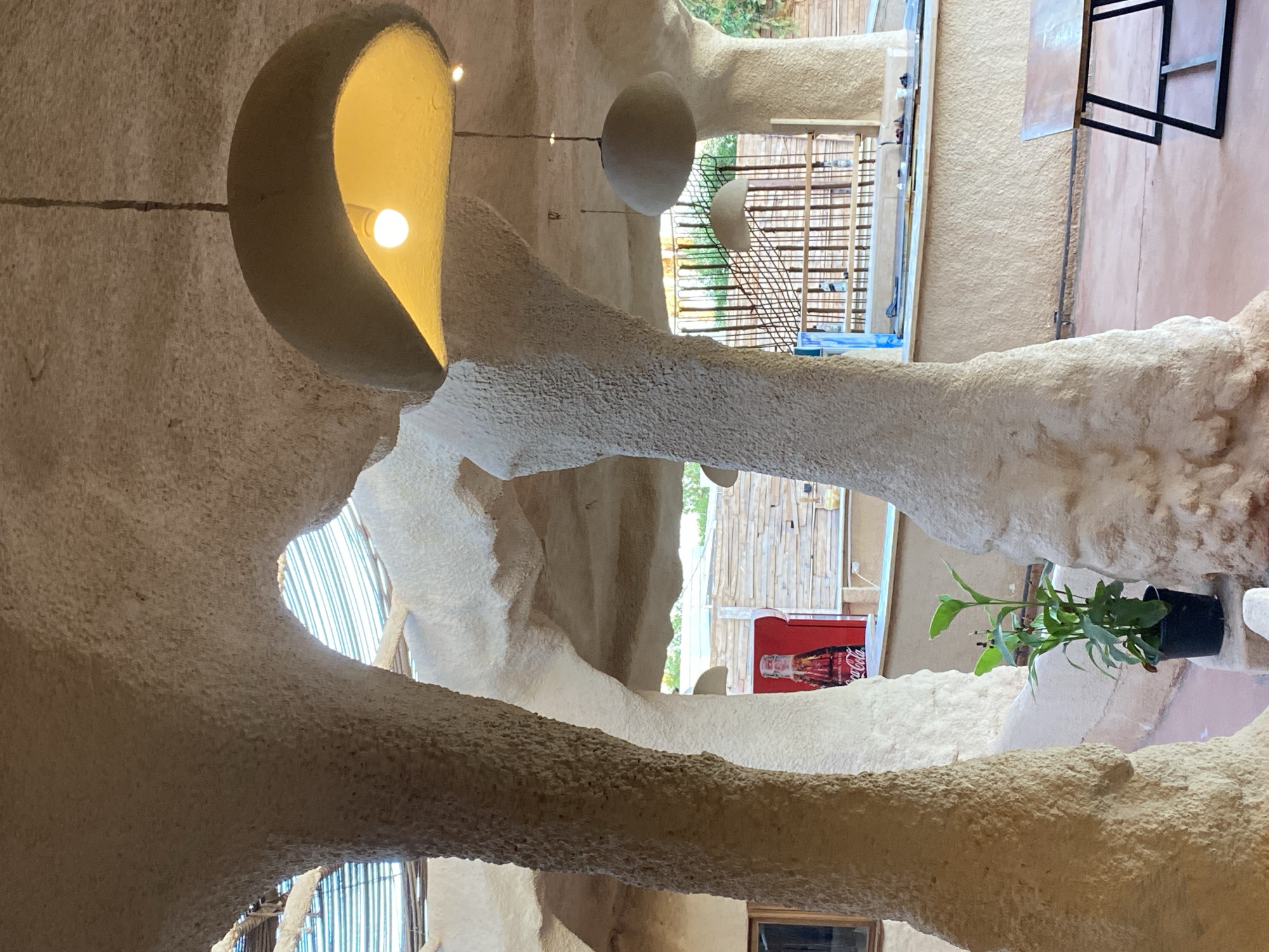 The image depicts the interior of a building with organic, cave-like architecture. The walls and structural elements are textured and appear to be made of plaster or a similar material, with smooth, curving forms reminiscent of natural rock formations. A bright, warm light emanates from a recessed fixture in the upper left, casting a soft glow. Other spherical light fixtures hang from the ceiling, adding to the soft, ambient lighting.
Through openings in the architecture, glimpses of the outside are visible. These include bamboo-like structures and greenery, suggesting an outdoor setting possibly in a warm climate. In one of these openings, a red Coca-Cola cooler with a classic bottle design is visible, along with visible text that reads "Coca-Cola". A small potted plant with green leaves is also placed in a recess in the wall, adding a touch of nature to the interior.
The overall impression is one of a unique and artistic space, possibly a cafe, restaurant, or lounge, designed to evoke a natural, earthy feel. The lighting is warm and inviting, and the organic shapes of the architecture create a sense of enclosure and comfort. There are no people visible in the image. The time of day is indeterminate, but the lighting suggests it is either daytime with filtered natural light or evening with artificial illumination. The location context of Sharm El Sheikh, Egypt, aligns with the potential use of natural materials and an aesthetic that might suit a resort or tourist area.
