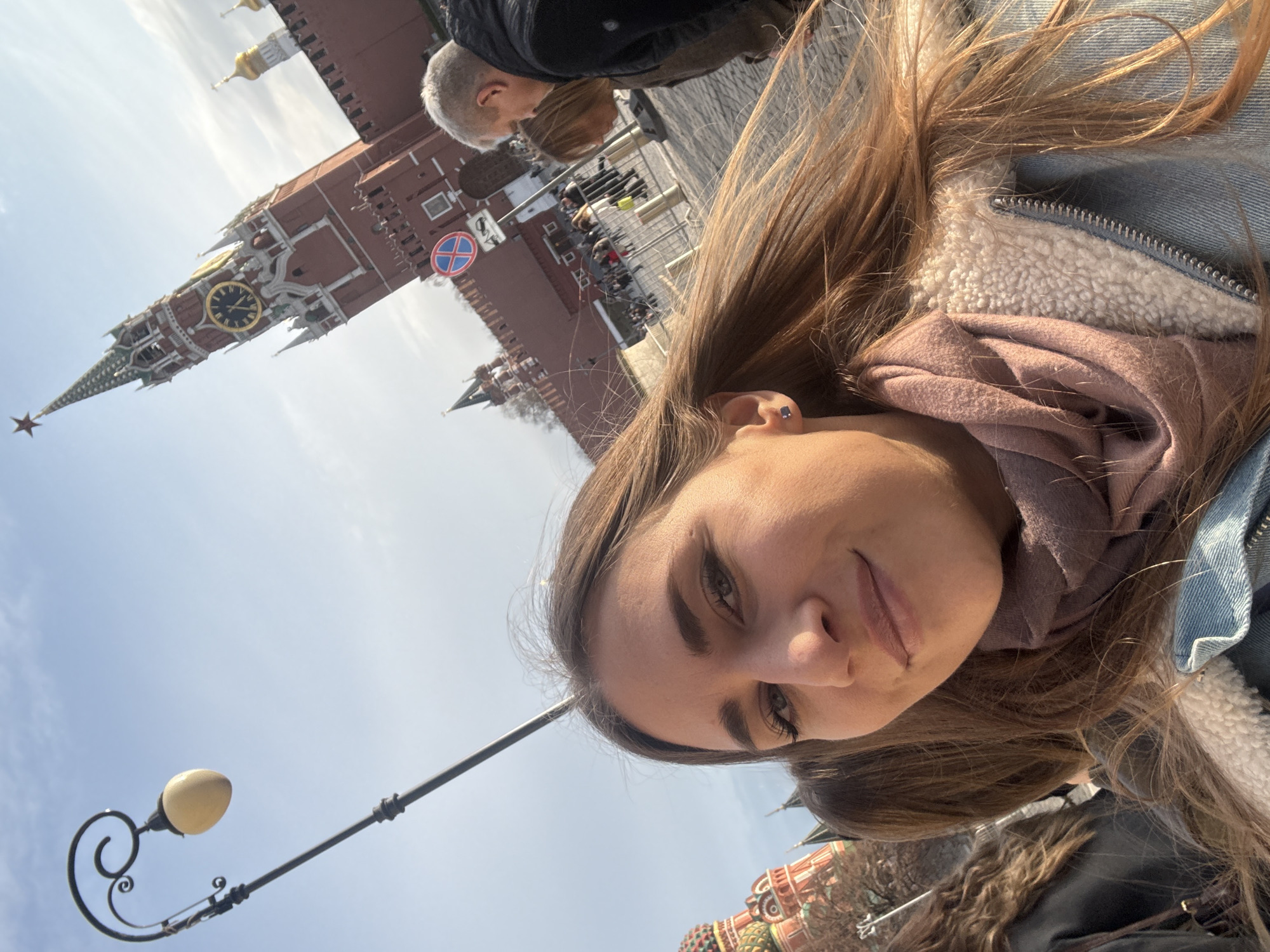The image depicts a daytime scene in Red Square, Moscow, Russia. In the immediate foreground, a young adult female with light brown hair is visible, looking directly at the camera with a slight smile, suggesting a selfie. She wears a light blue denim jacket with a white sherpa lining and a pink scarf around her neck. A small dark stud earring is visible in her left ear. To her left, part of another individual's head with brown hair is visible.
To the left of the main subject, a black ornamental street lamp with a yellowish-white spherical light fixture extends upwards. In the midground, the scene features a cobblestone pavement, metal crowd barriers, and a blue circular "No Stopping" sign (red cross on blue background) on a pole. Several indistinct figures are visible beyond the barriers, including a man with gray hair in a dark jacket, partially obscured and facing away from the camera.
The background prominently displays the Spasskaya Tower of the Moscow Kremlin, a red brick structure with white ornamentation, a golden clock face, and a green spire topped with a red star. A section of the red brick Kremlin wall is also visible. On the far left, the distinct multi-colored onion domes of St. Basil's Cathedral are apparent. The sky is clear blue with minimal cloud cover, and bright sunlight illuminates the scene.