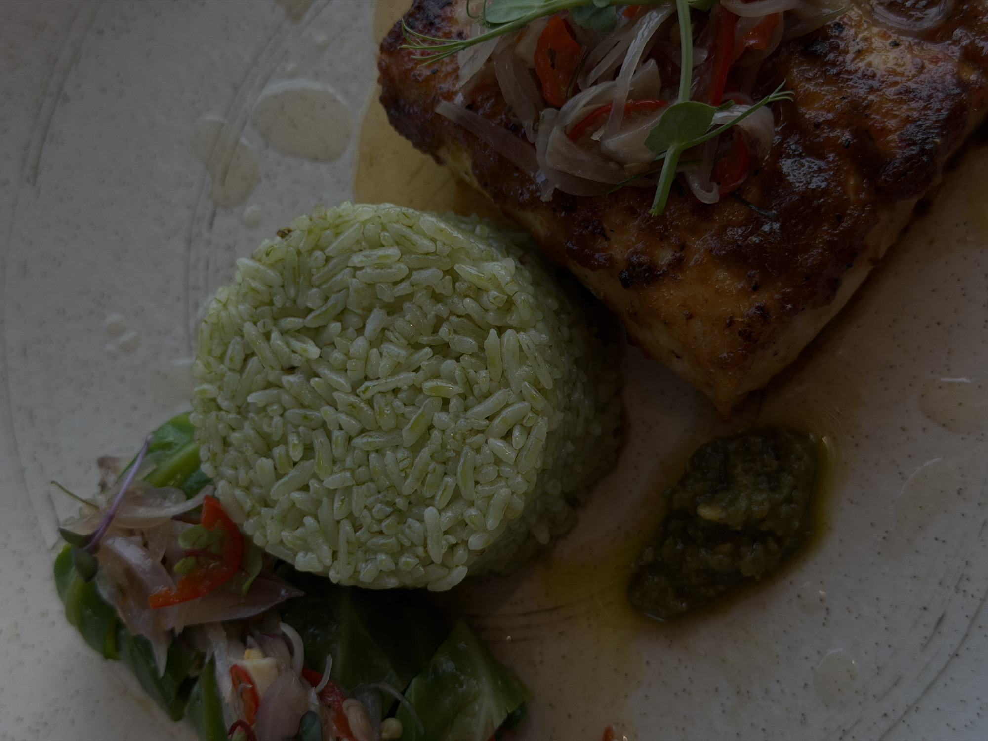 A close-up, overhead shot displays a plated meal on a textured, light-colored ceramic dish in Belalang, Indonesia. The centerpiece is a golden-brown piece of seared fish or poultry, topped with a relish composed of thinly sliced shallots, red chili, and fresh green microgreens. To the left of the main protein is a cylindrical mound of light green-colored rice. Further to the upper left, a portion of green leafy salad, intermixed with sliced shallots and red chili, is visible. A small dollop of dark green, textured sauce or paste is present on the lower left of the plate. The plate exhibits several oil or sauce streaks and speckling. No individuals are present in the frame.
